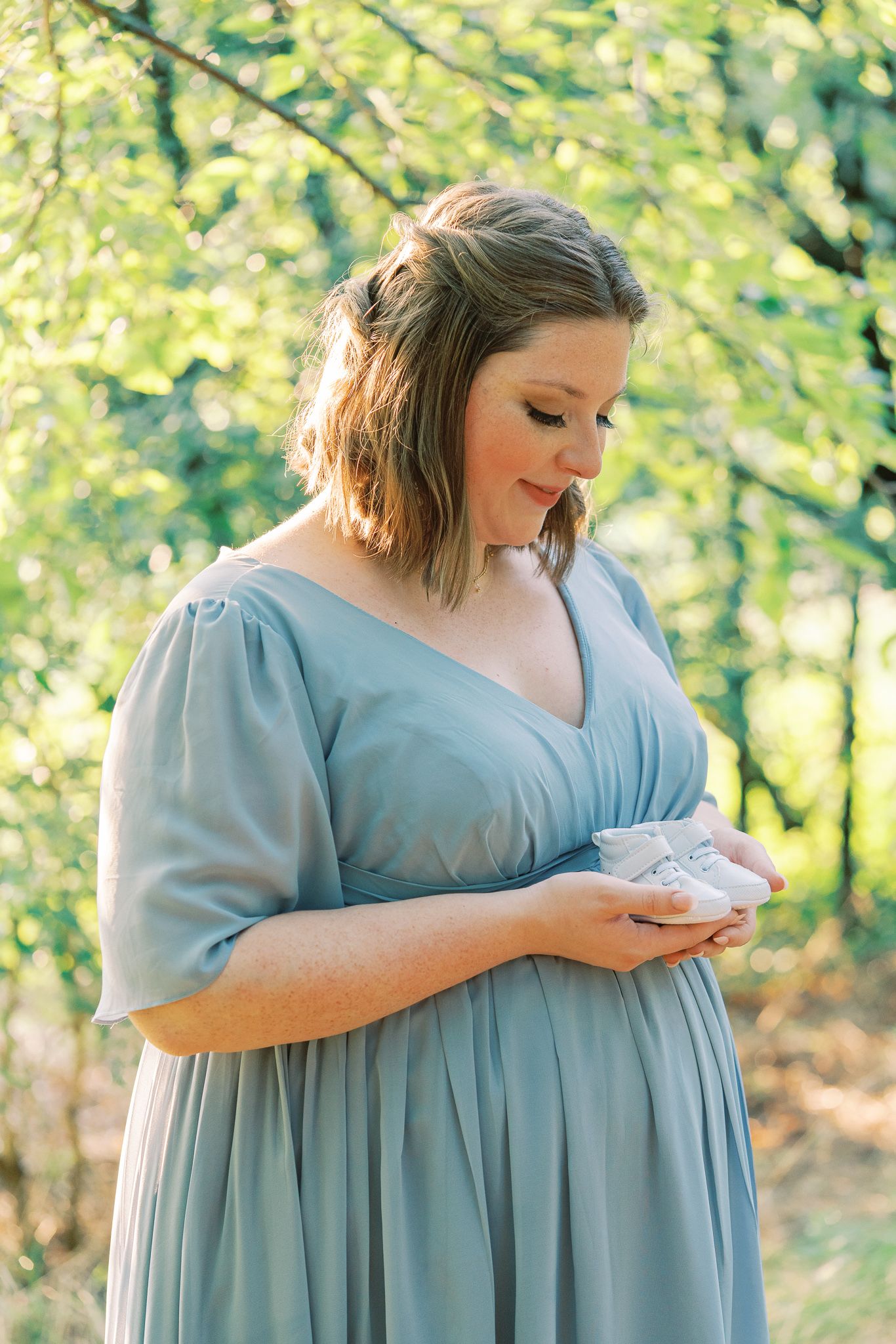 Expectant mother in a flowing blue dress stands in sunlit forest setting while looking down at her baby bump.