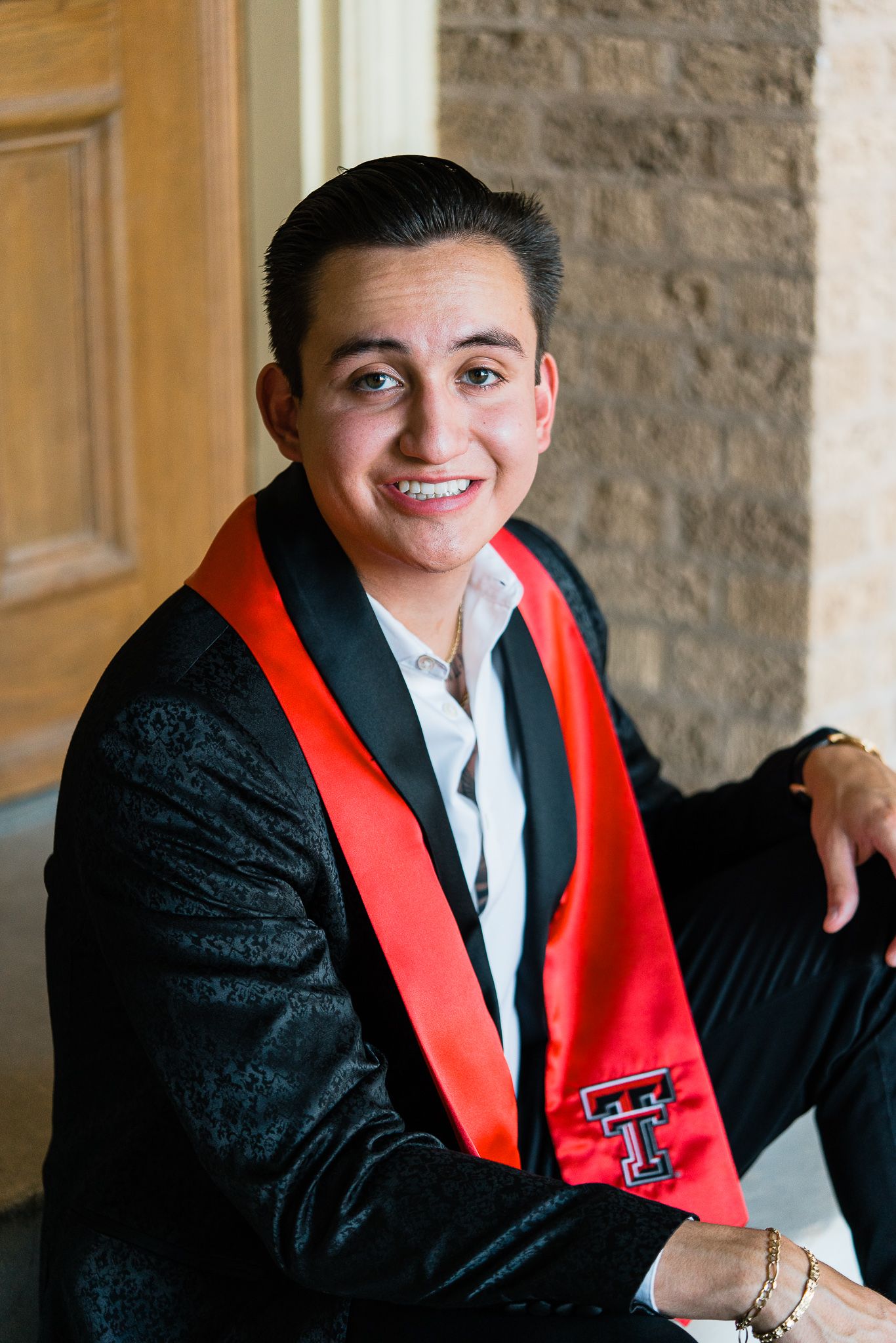 Graduate in black gown and red stole poses for a series of professional portraits against stone wall background.