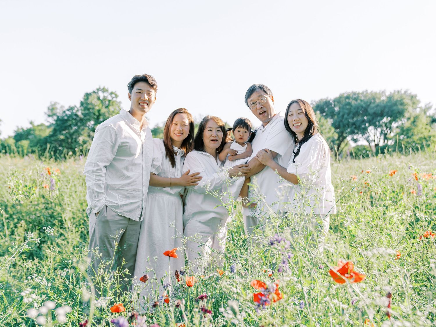 Group wearing white outfits poses joyfully in a blooming wildflower field on a sunny day.