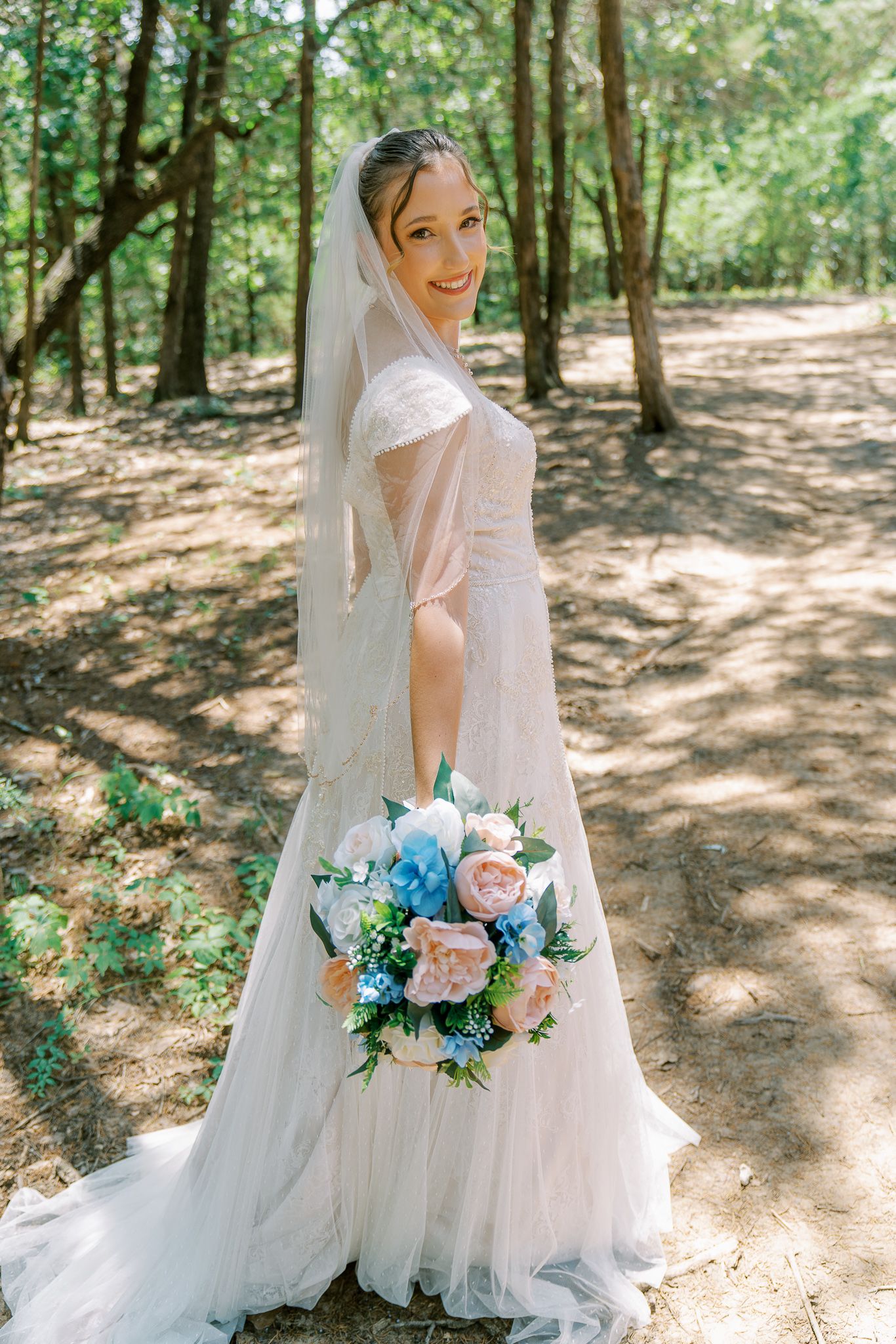 A bride in a flowing white gown poses with her bouquet featuring pink and blue flowers in a sunlit forest.