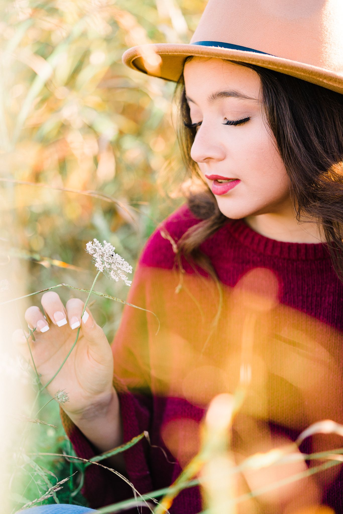 A woman in a burgundy sweater and brown hat poses in autumn foliage with soft bokeh lighting effects.