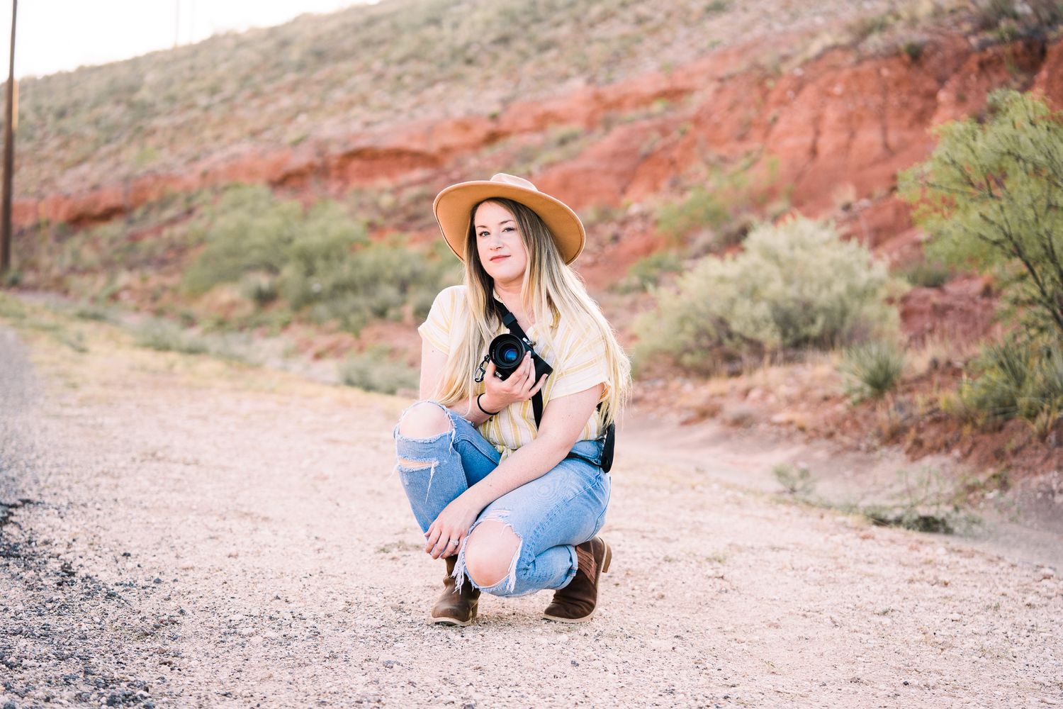 Woman in wide brim hat posing on desert trail with red rock formations in background.