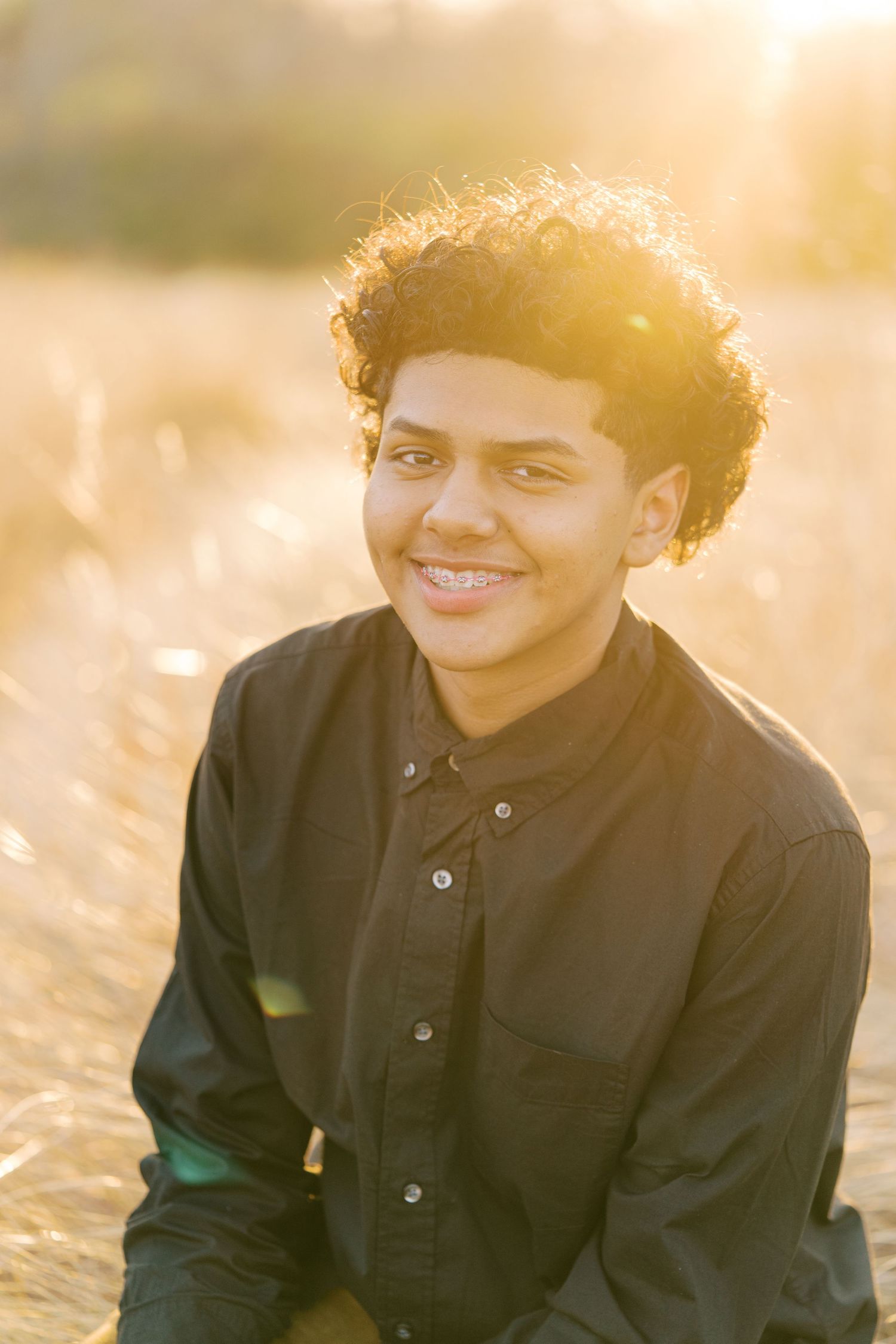 Person wearing a black button-up shirt smiles warmly in golden sunlight during an outdoor portrait session.