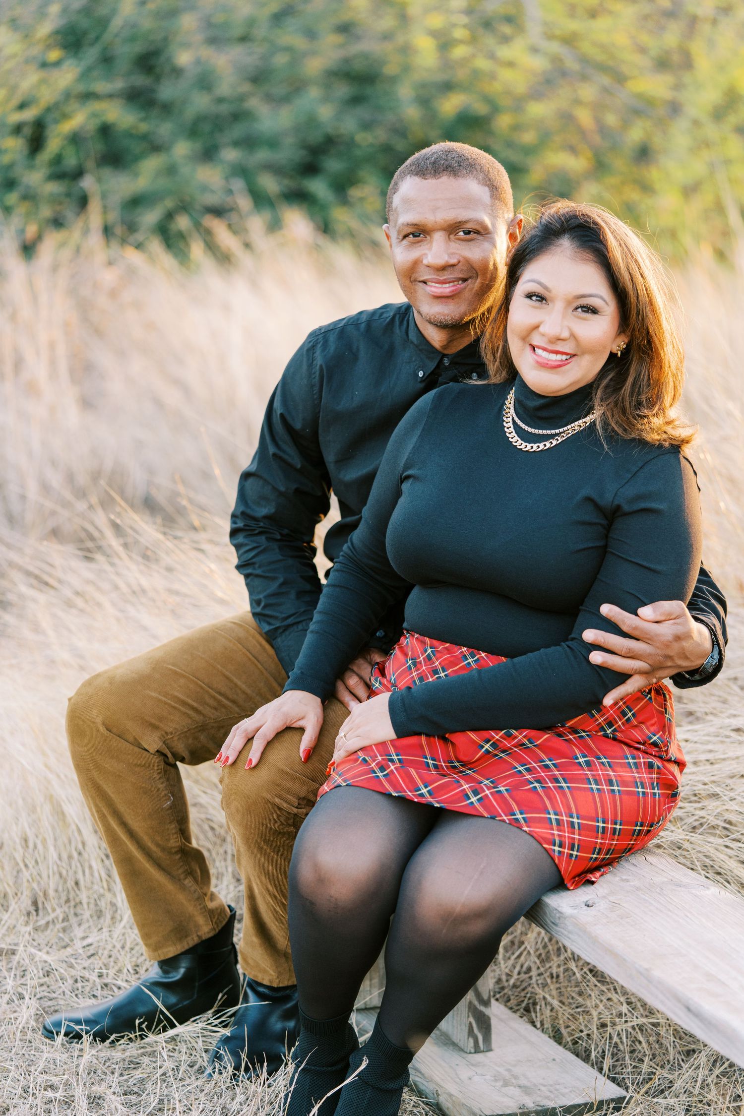Couple sits together on ground amid tall grass during sunset photoshoot wearing coordinated black and plaid outfits.