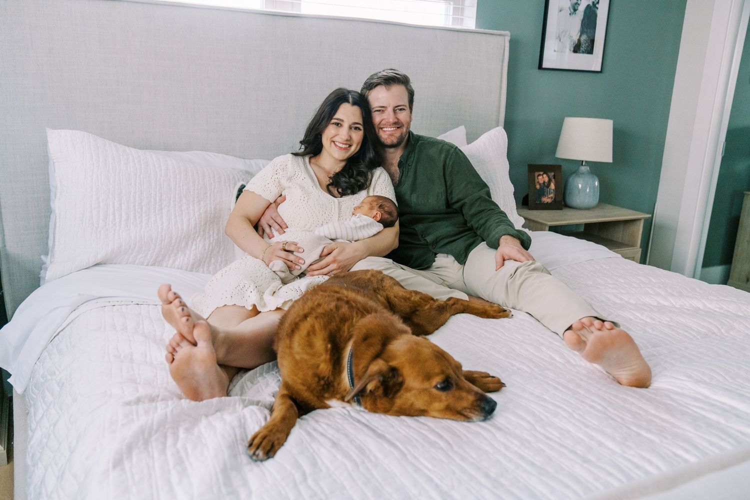 A couple and their dog relax together on a white bed in a cozy bedroom with teal walls.