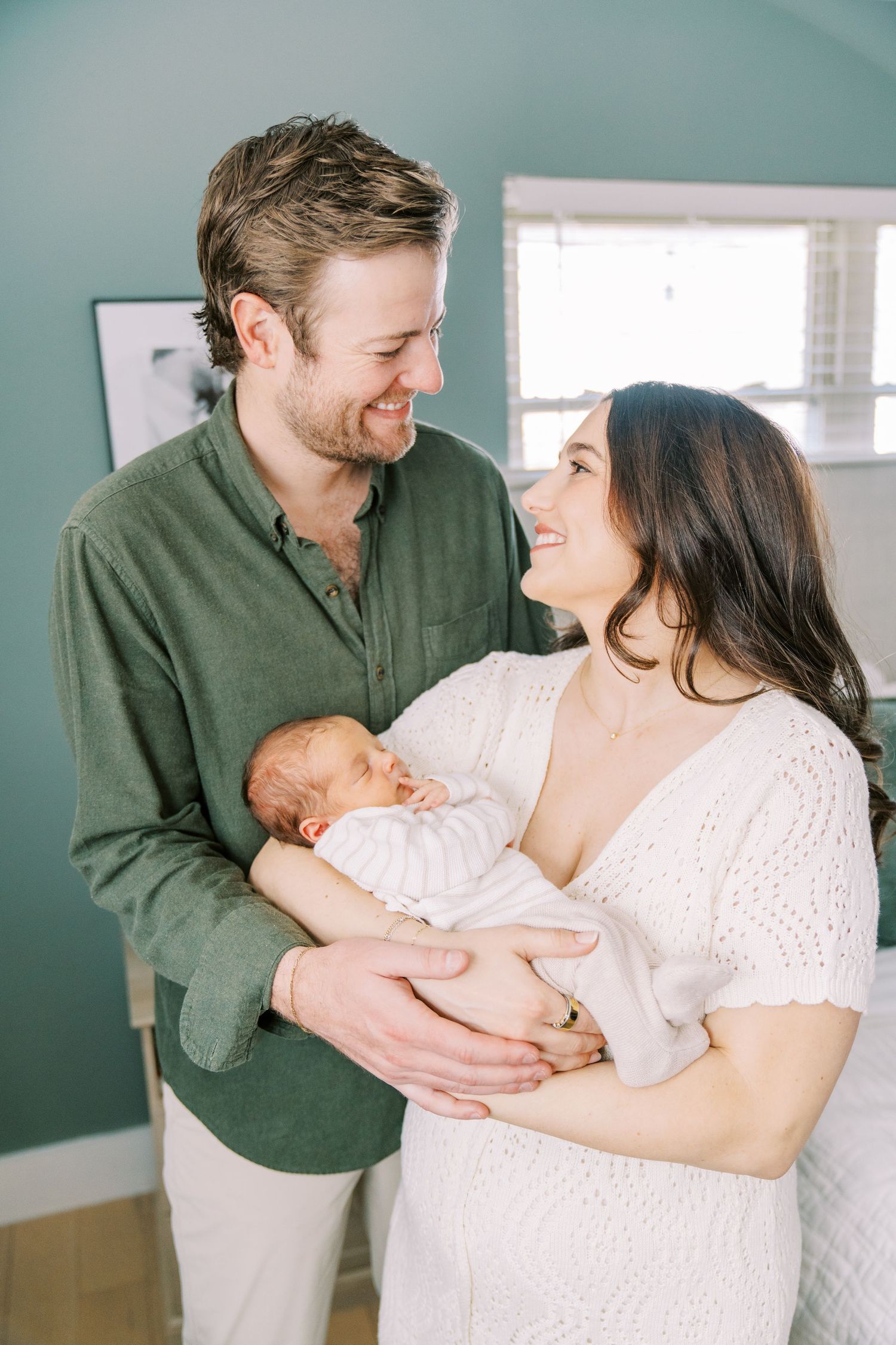 Happy couple in casual attire smiling while holding their newborn baby in a room with teal walls.
