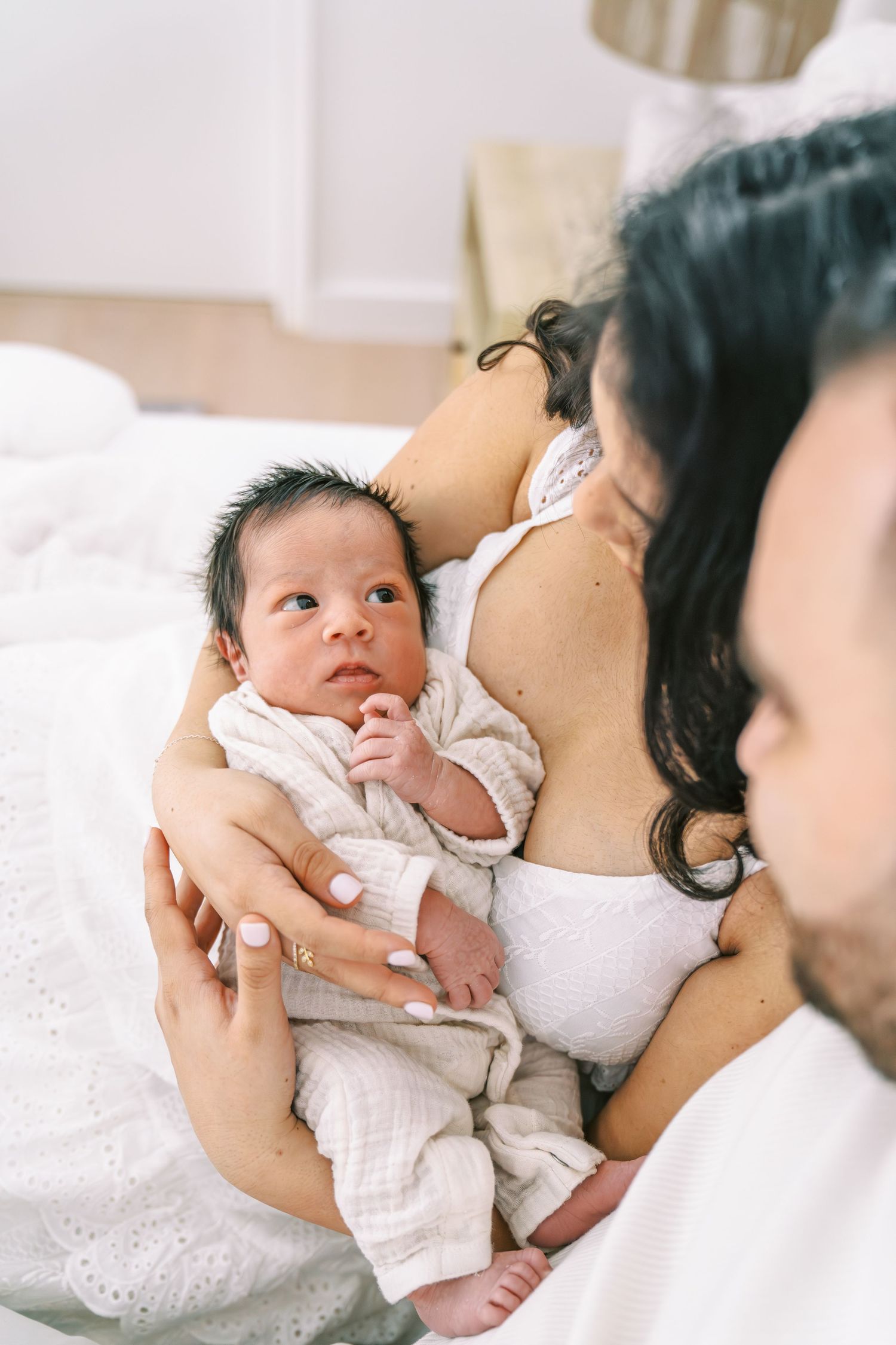 Parents cradling their newborn baby wrapped in a white muslin swaddle on a bed.