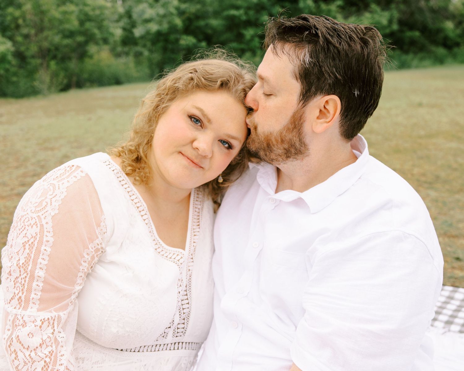 A man gently kisses a smiling woman's cheek as they sit together on a blanket in a green park.