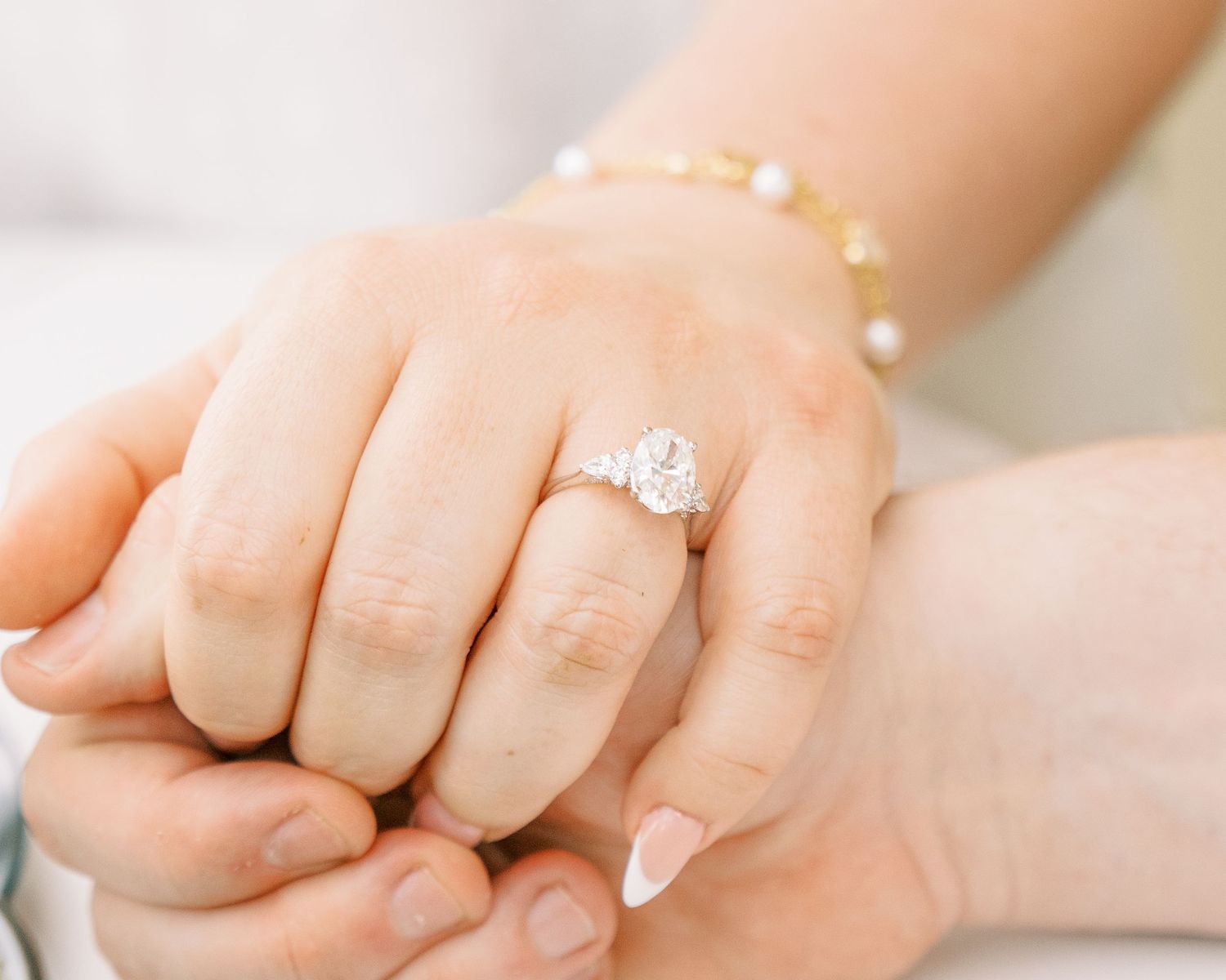 A close-up of an elegant oval diamond engagement ring on a woman's hand with a pearl bracelet.