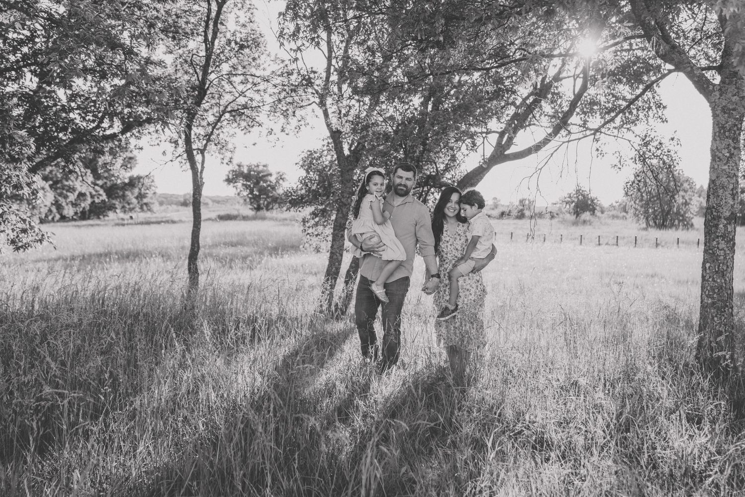 Black and white photo of a family walking through a sunlit field surrounded by trees on a warm day.