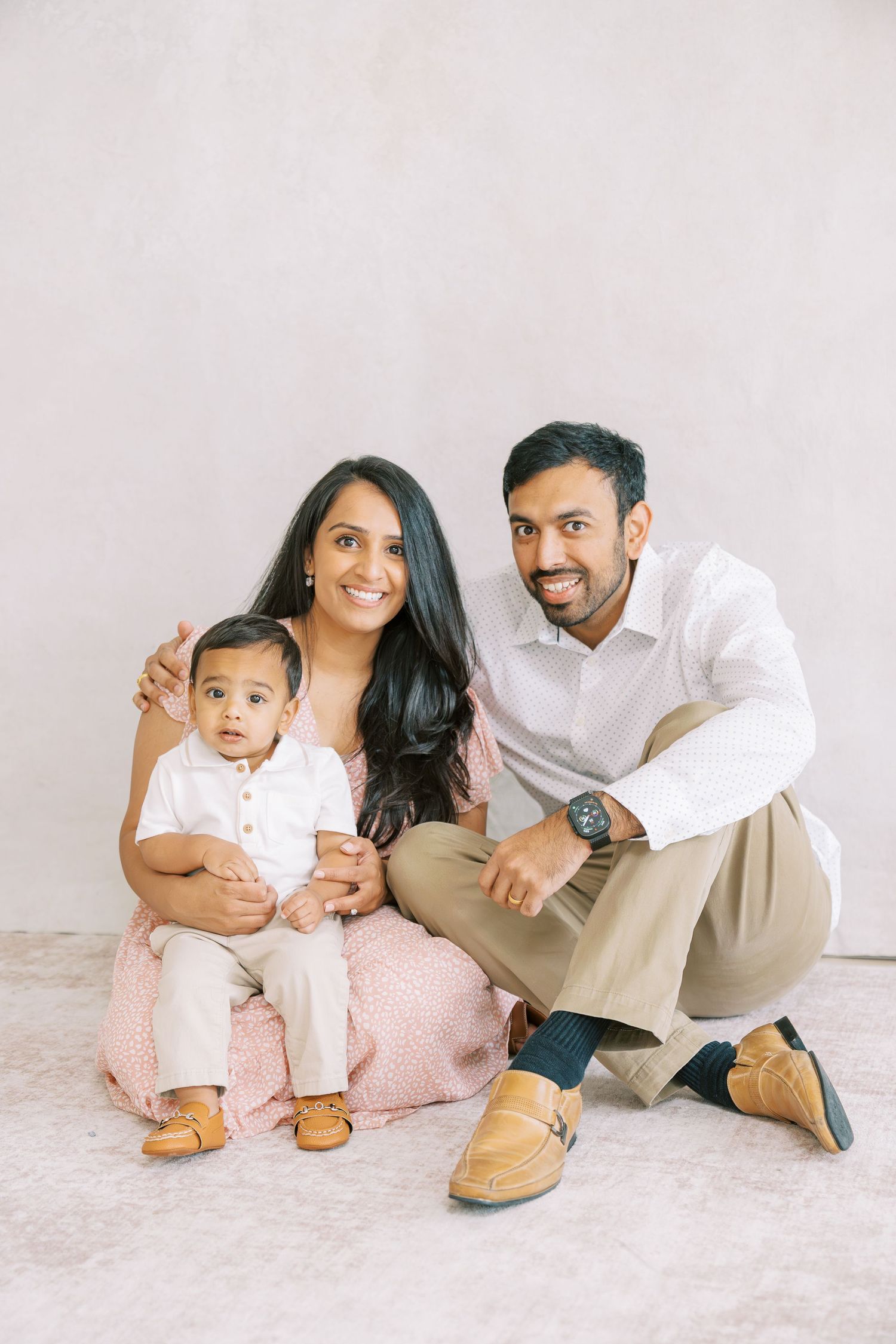 A happy South Asian family with a baby boy poses together on the floor in coordinating neutral outfits for a studio portrait.