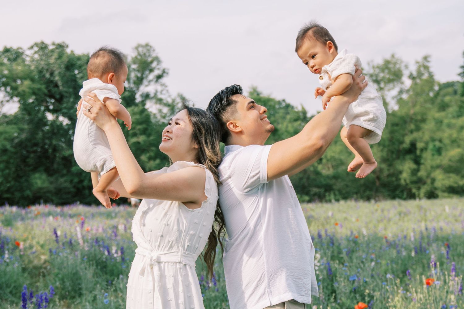 Happy Asian family in white outfits lifting twin babies in a colorful wildflower meadow on a sunny day.