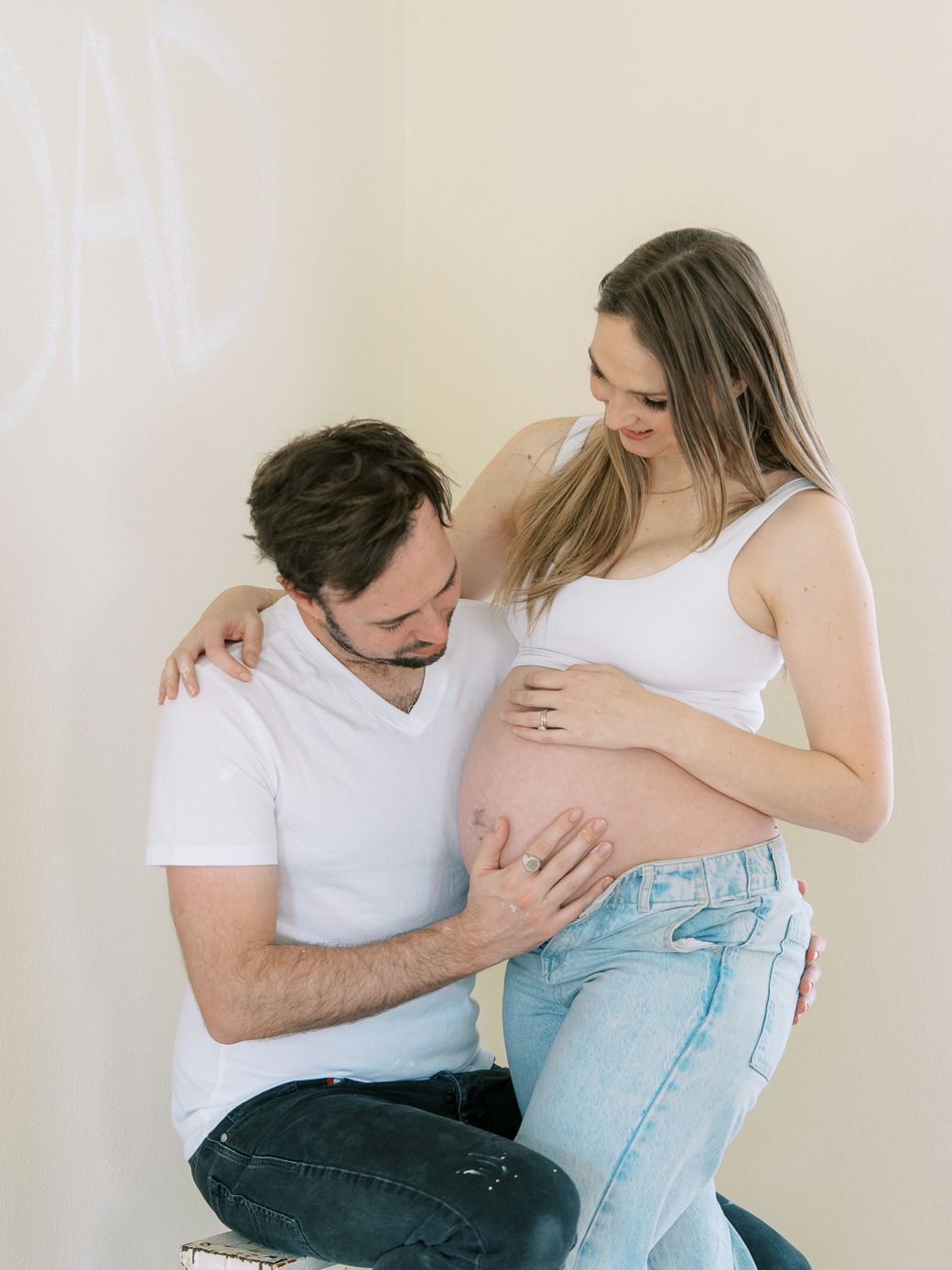 Maternity photoshoot showing intimate moments between expectant parents in white clothing against neutral background.