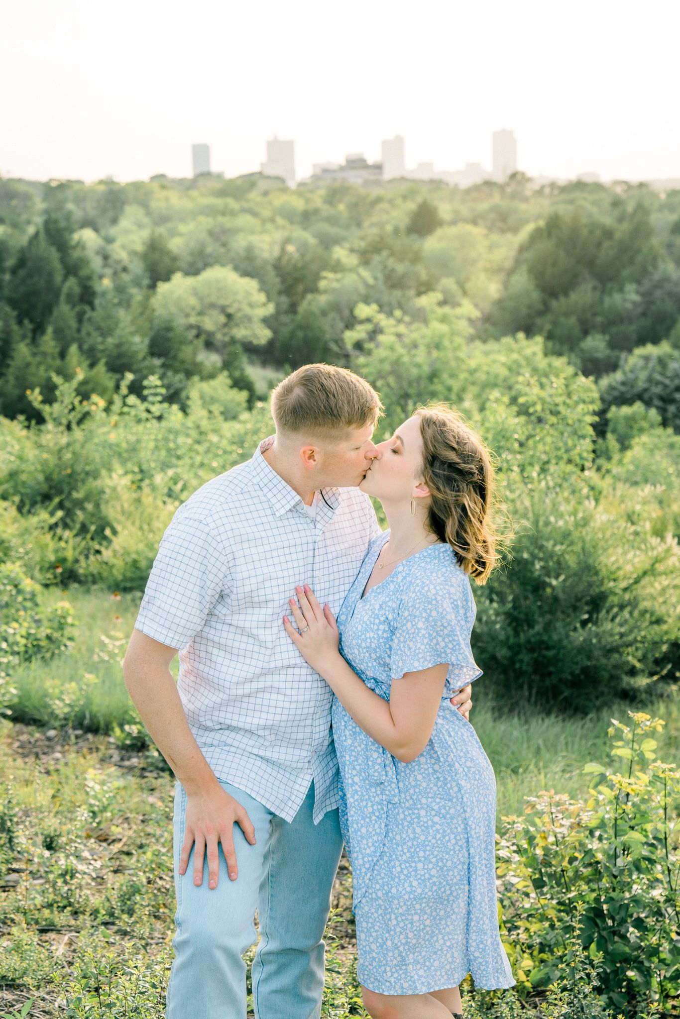 Romantic couple in blue outfits embracing in a sunny meadow with city skyline in background.