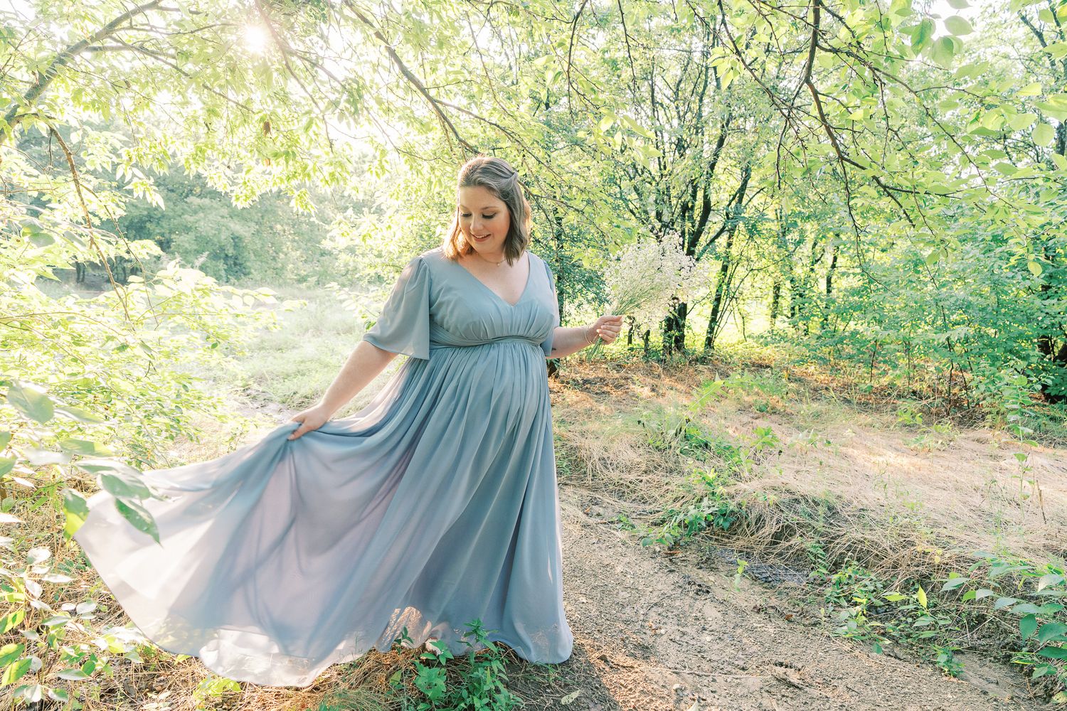 A person in a flowing dusty blue maxi dress twirls on a sunlit forest path during golden hour.