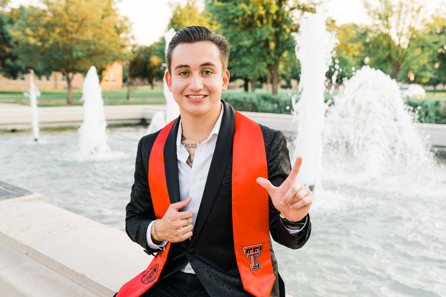 Graduate in black suit and red stole poses near outdoor fountain during graduation photo session.