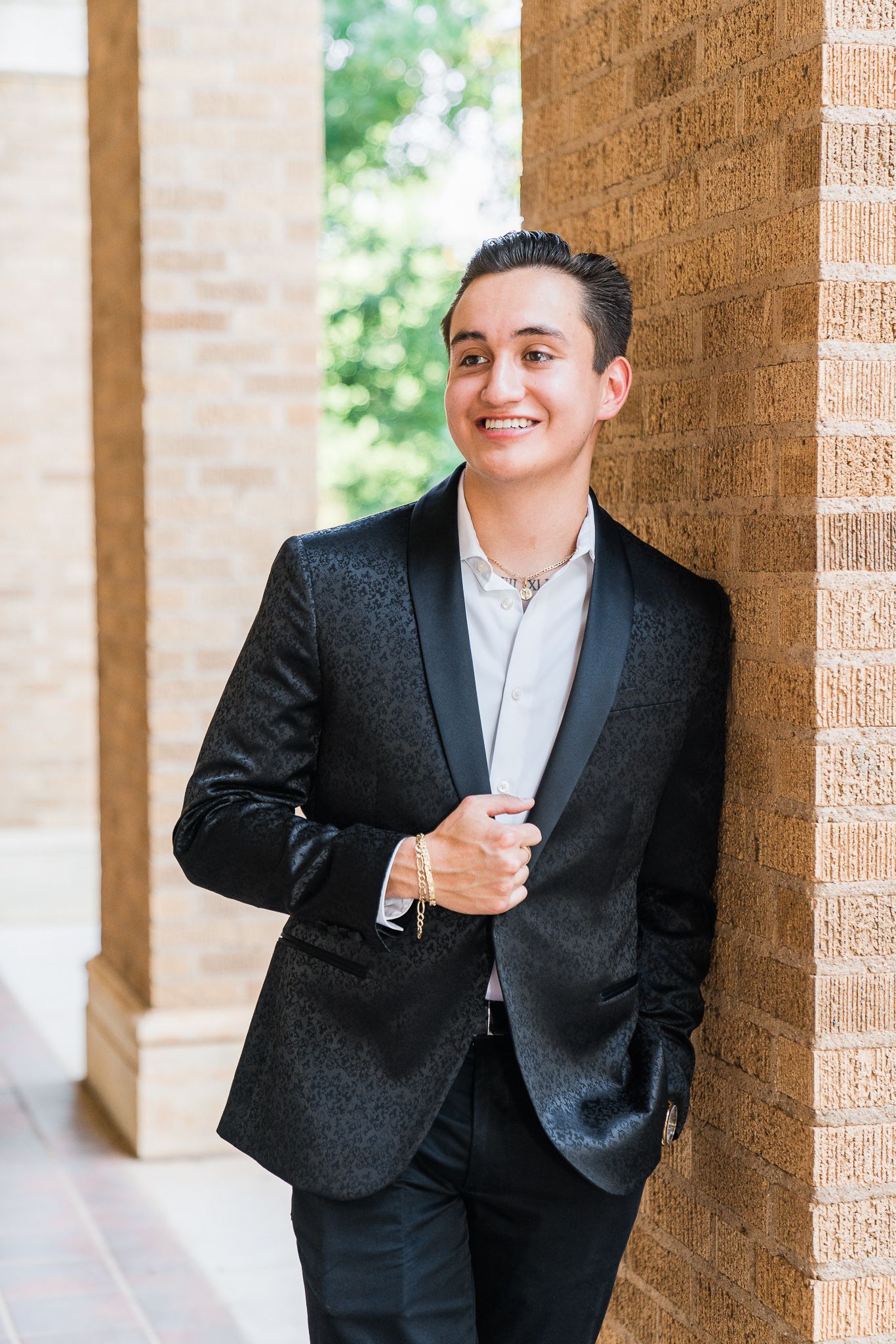 A person in a black suit and white shirt poses cheerfully against a brick wall with columns.
