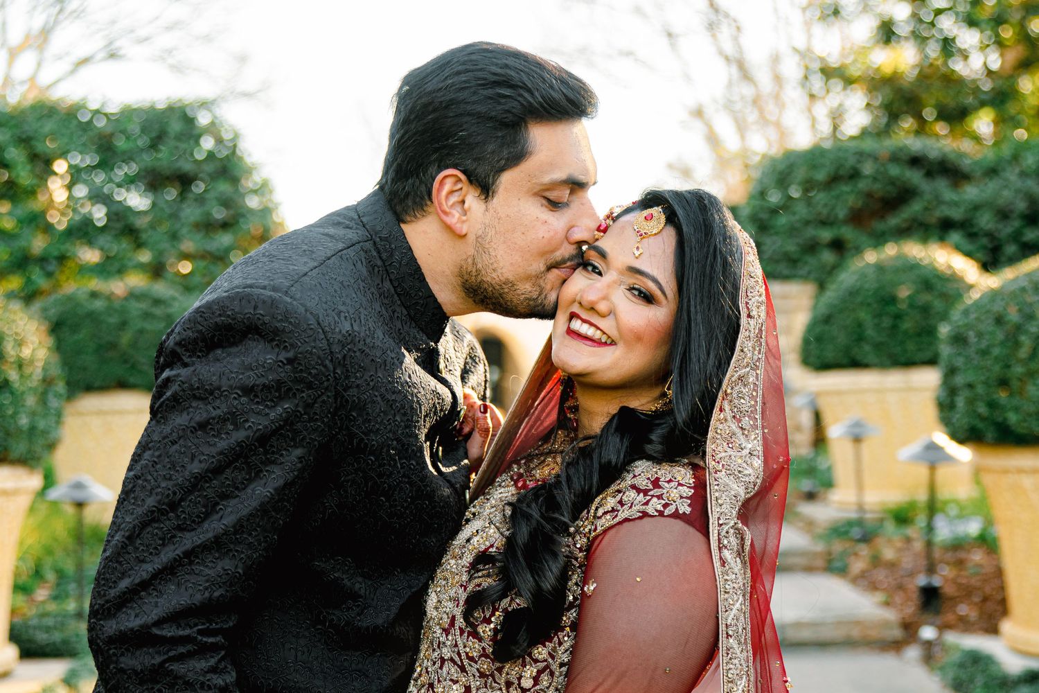 A romantic couple in traditional South Asian wedding attire share intimate moments in an outdoor garden setting.
