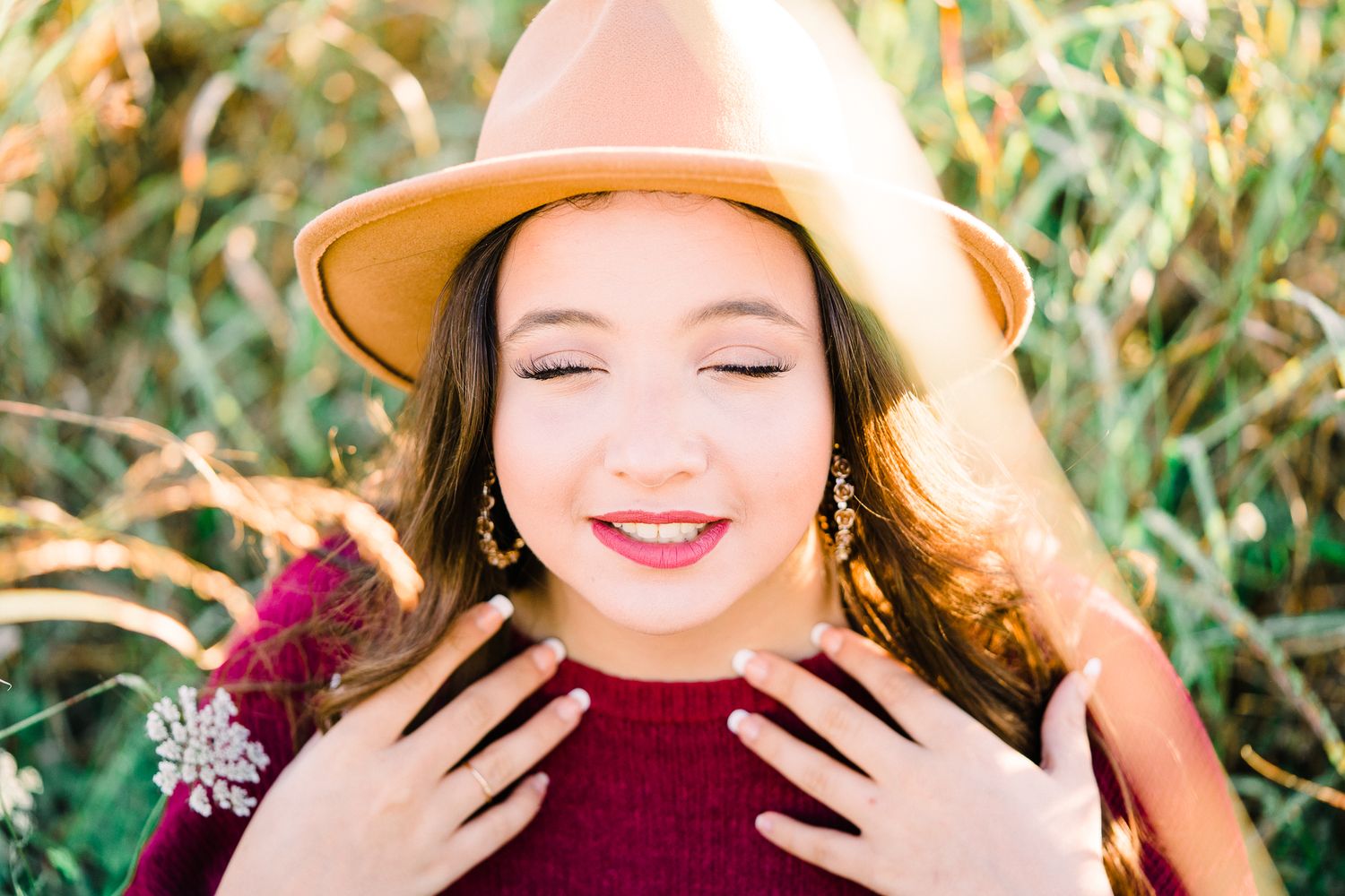 A person wearing a burgundy sweater and brown hat poses outdoors in autumn with greenery in the background.