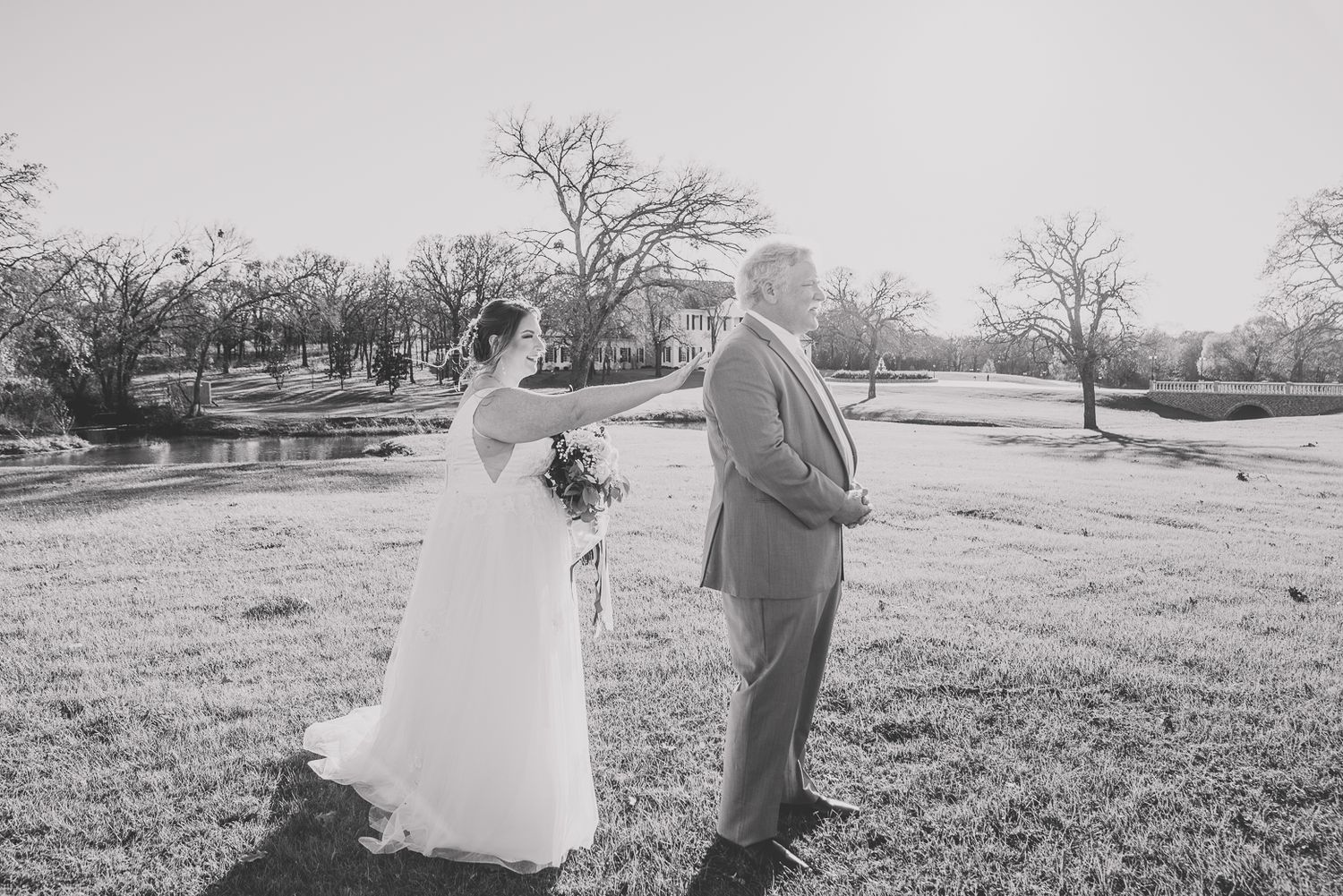 A romantic couple in wedding attire dances and spins together in a sunny outdoor field.