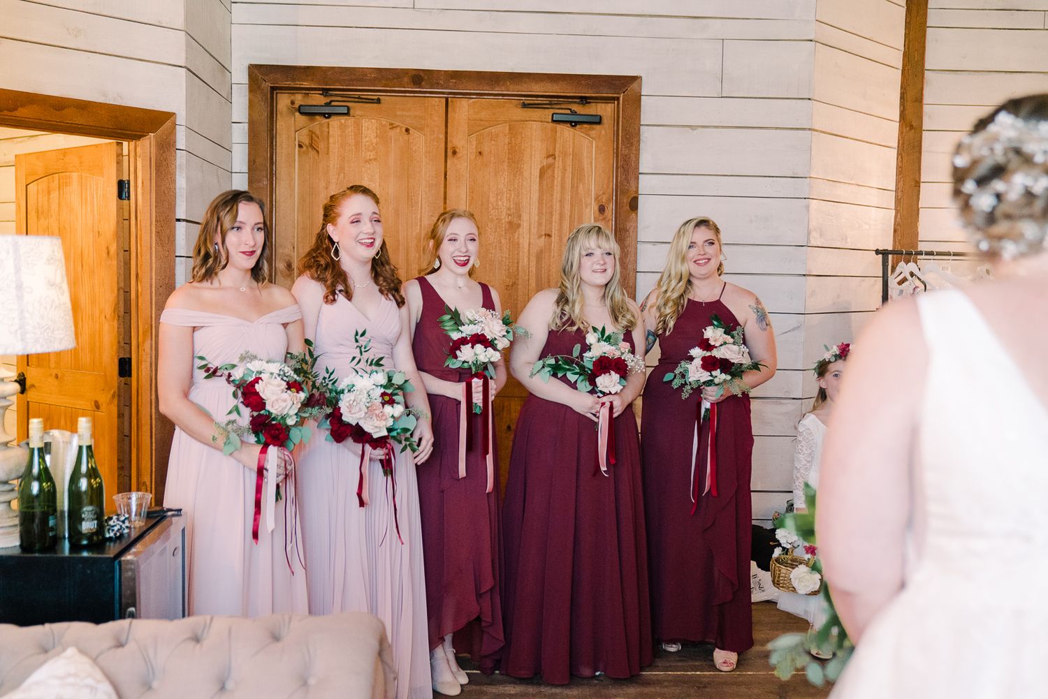 Wedding party members in burgundy and blush dresses holding coordinated floral bouquets inside a rustic venue.