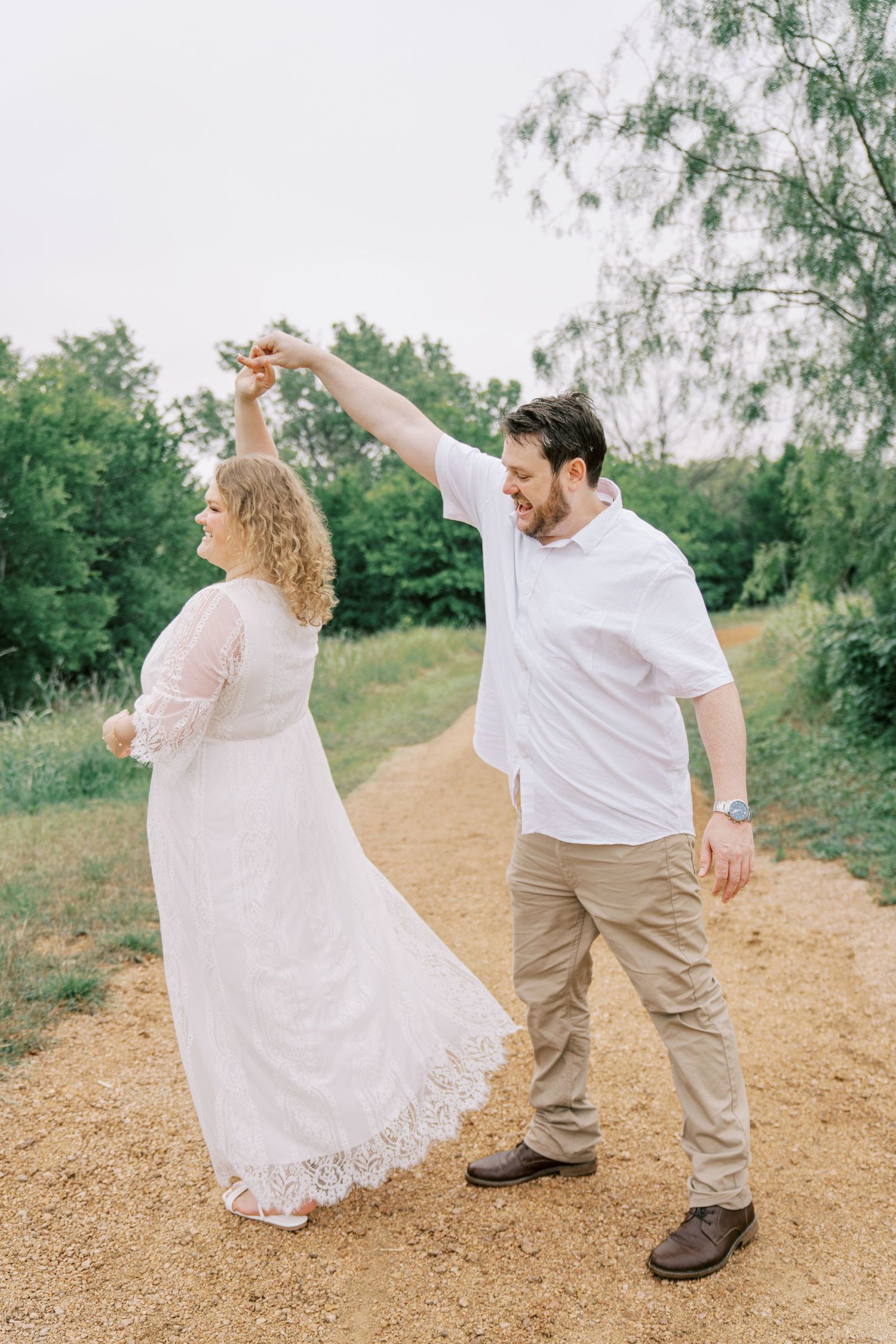 Joyful couple dancing on a dirt path outdoors, man in white shirt spinning woman in flowing white lace dress.