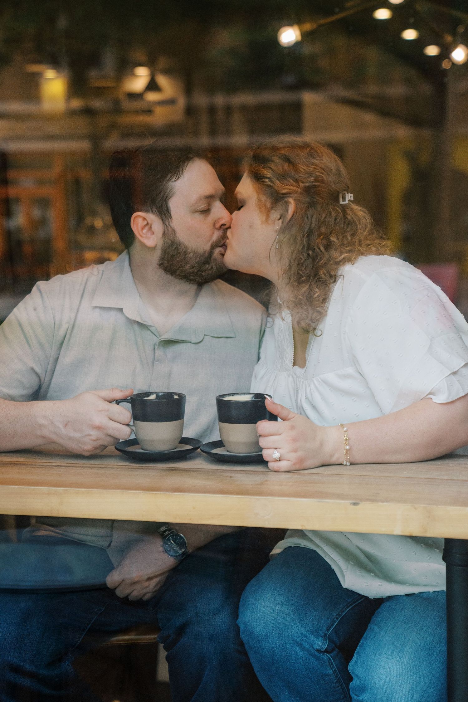 Couple sharing a romantic kiss over coffee cups at a cozy cafe table, viewed through a window.