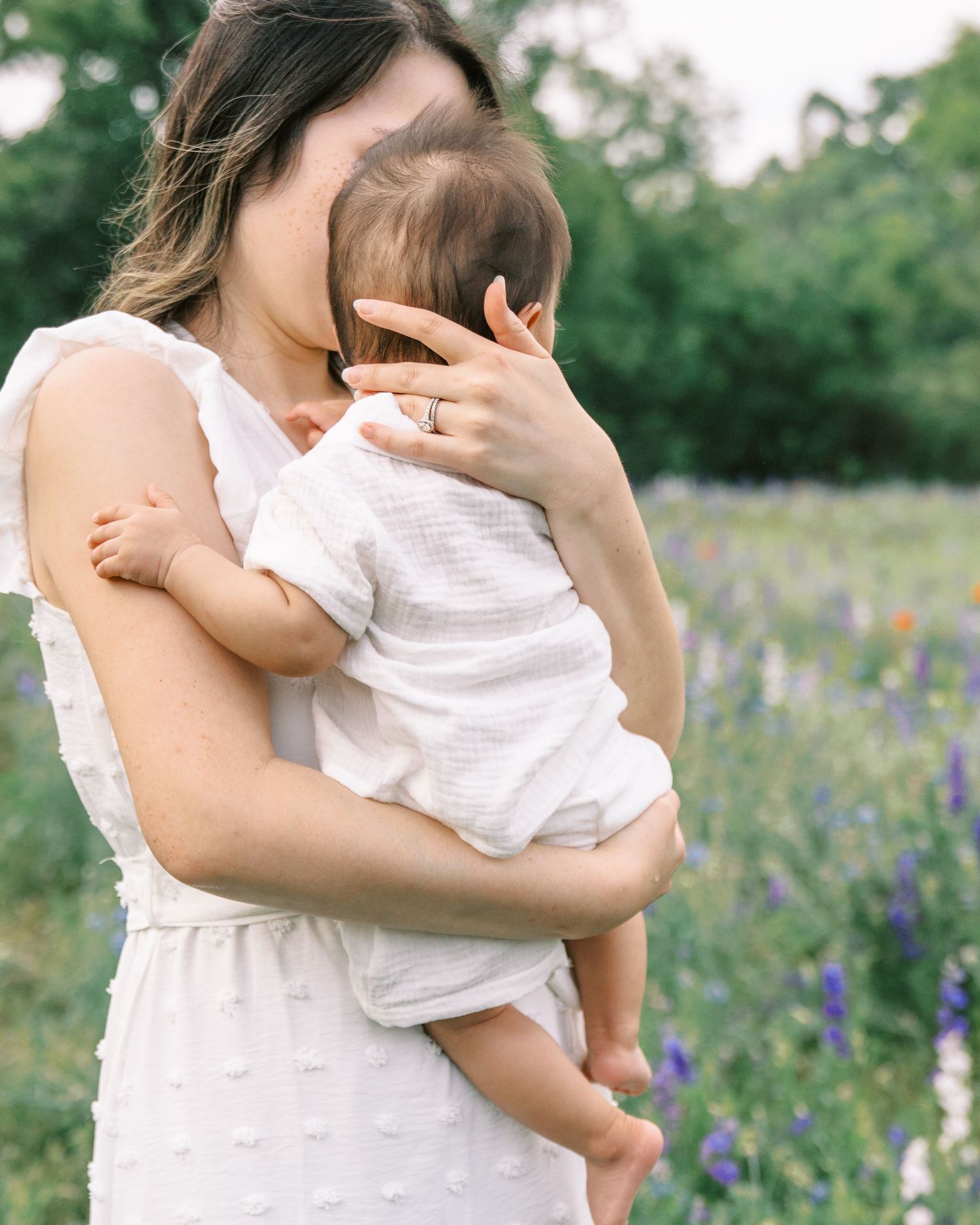 A mother in a white dress holds her baby in a wildflower field, gently kissing the infant's head.
