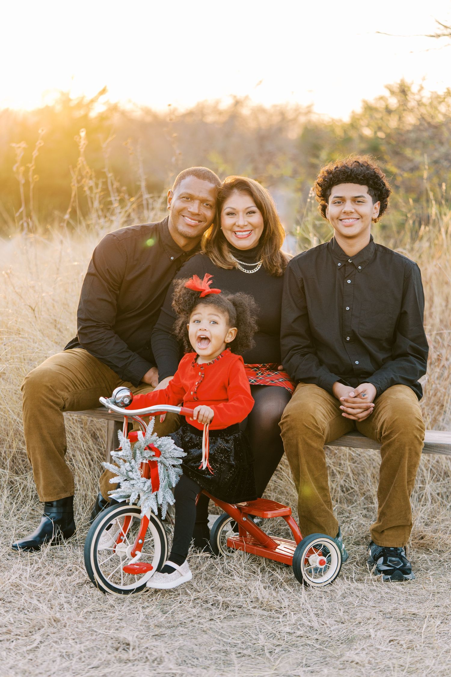 Family of four posing outdoors at golden hour, with a little girl on a red tricycle, all wearing black and red outfits.