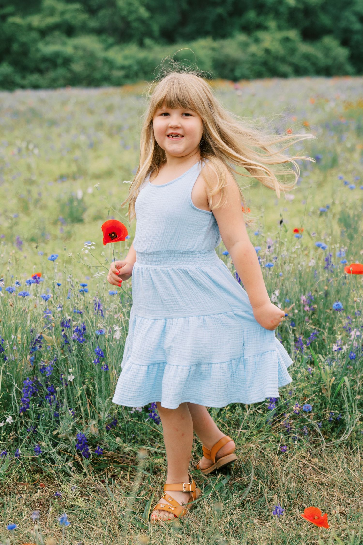 Young blonde girl in blue dress holding red poppy flower in colorful wildflower meadow, smiling joyfully.