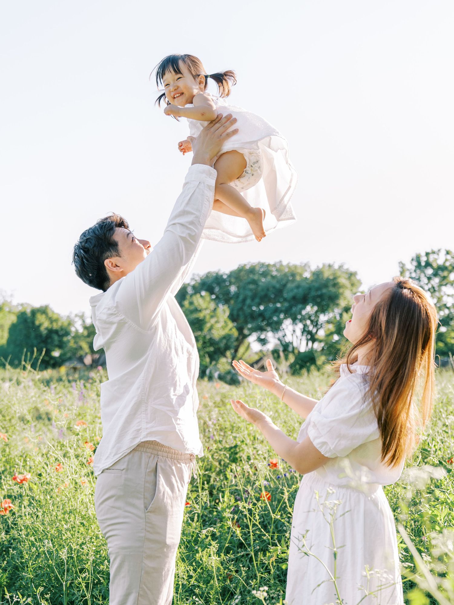 A joyful family in white outfits plays in a sunny meadow, father lifting smiling toddler girl while mother watches.