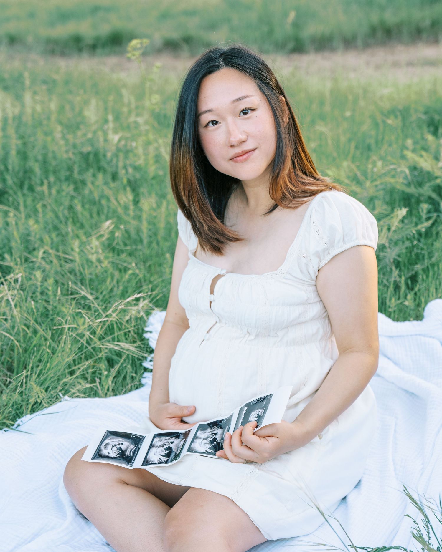 Pregnant woman in white dress sitting in a field holding ultrasound photos during a maternity photoshoot.