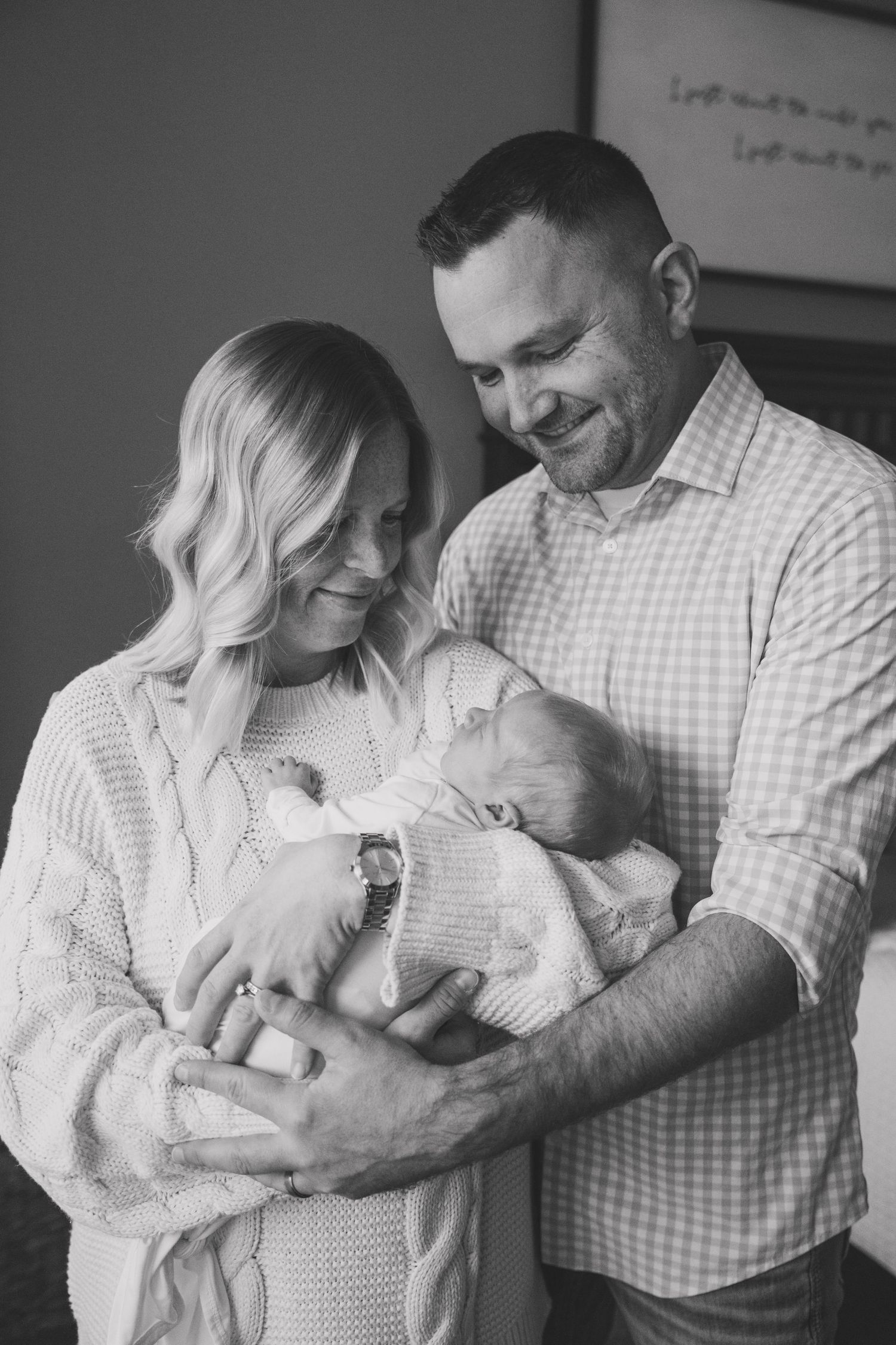 Parents in matching white outfits tenderly hold and gaze at their newborn baby in a series of intimate black and white photos.