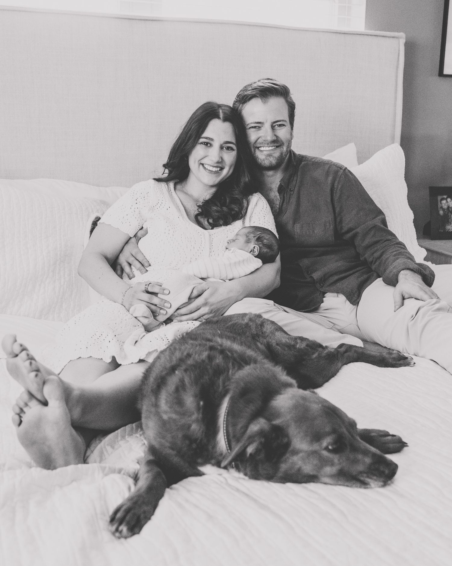 Black and white photo showing a couple relaxing on a bed with a newborn baby and a large sleeping dog.