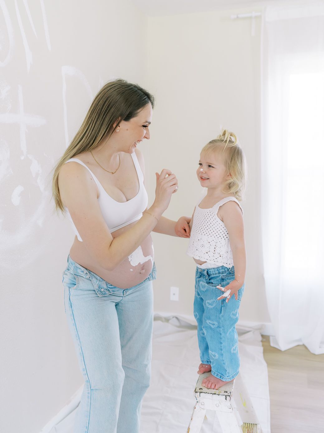 A parent and toddler share a playful moment in matching white tops and light denim jeans against a bright white backdrop.