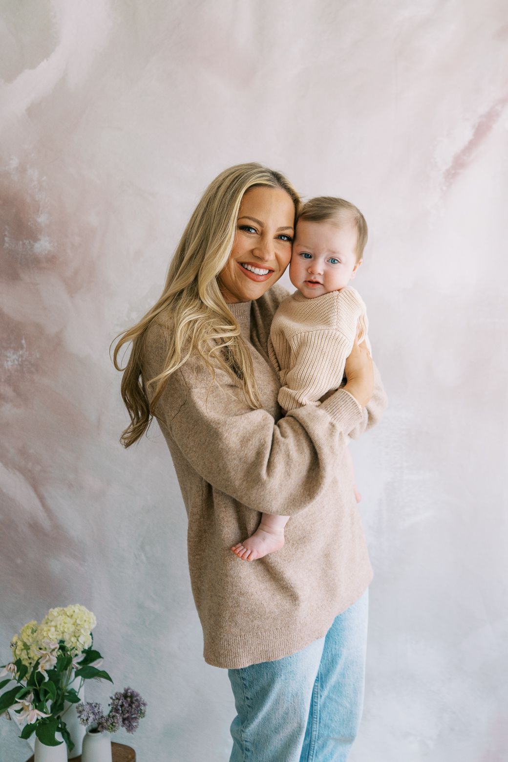 Mother and baby sharing tender moments near floral arrangement against light wall.