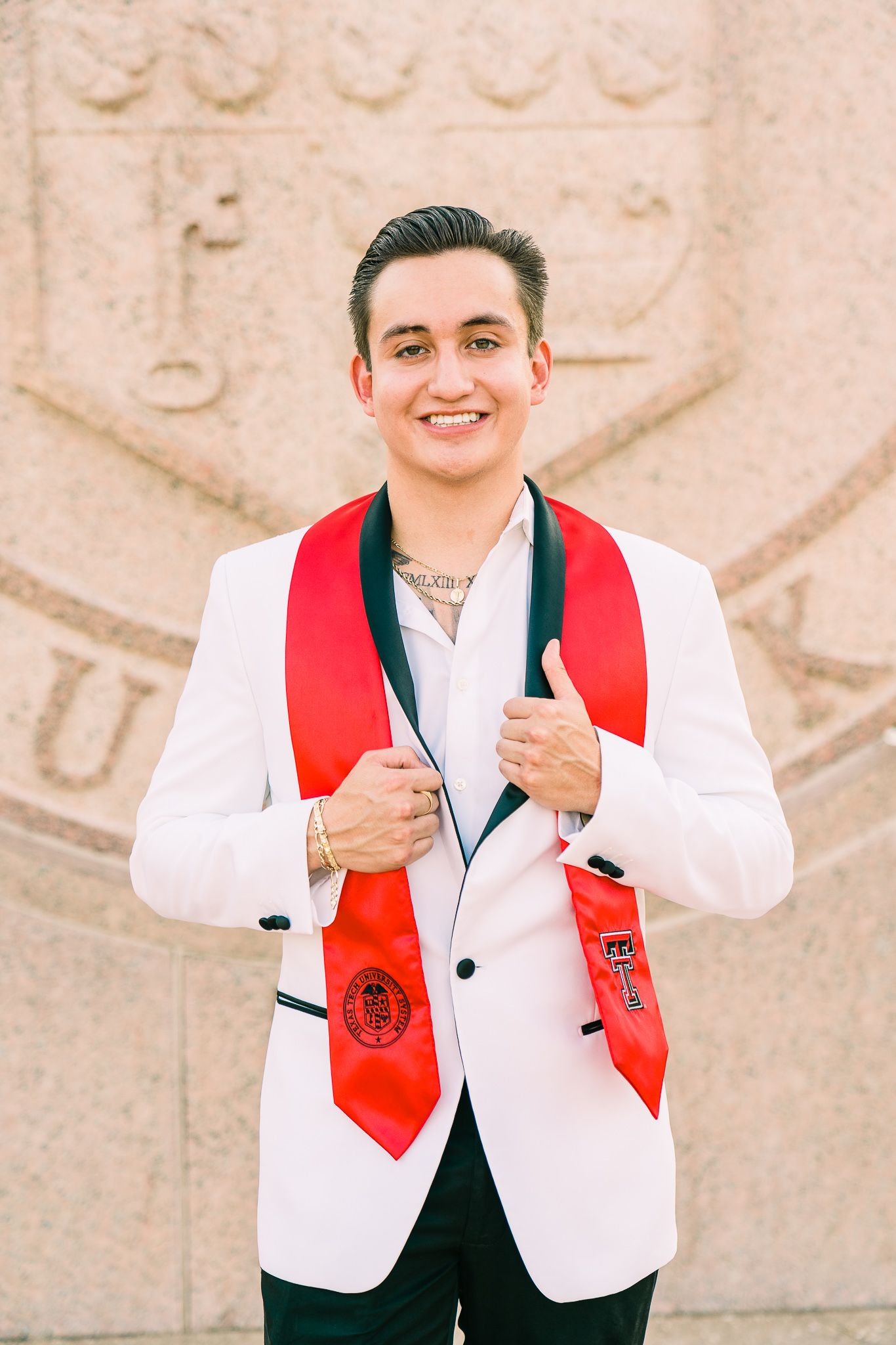 Graduate in white jacket and red stole gestures while standing against a decorative stone background.