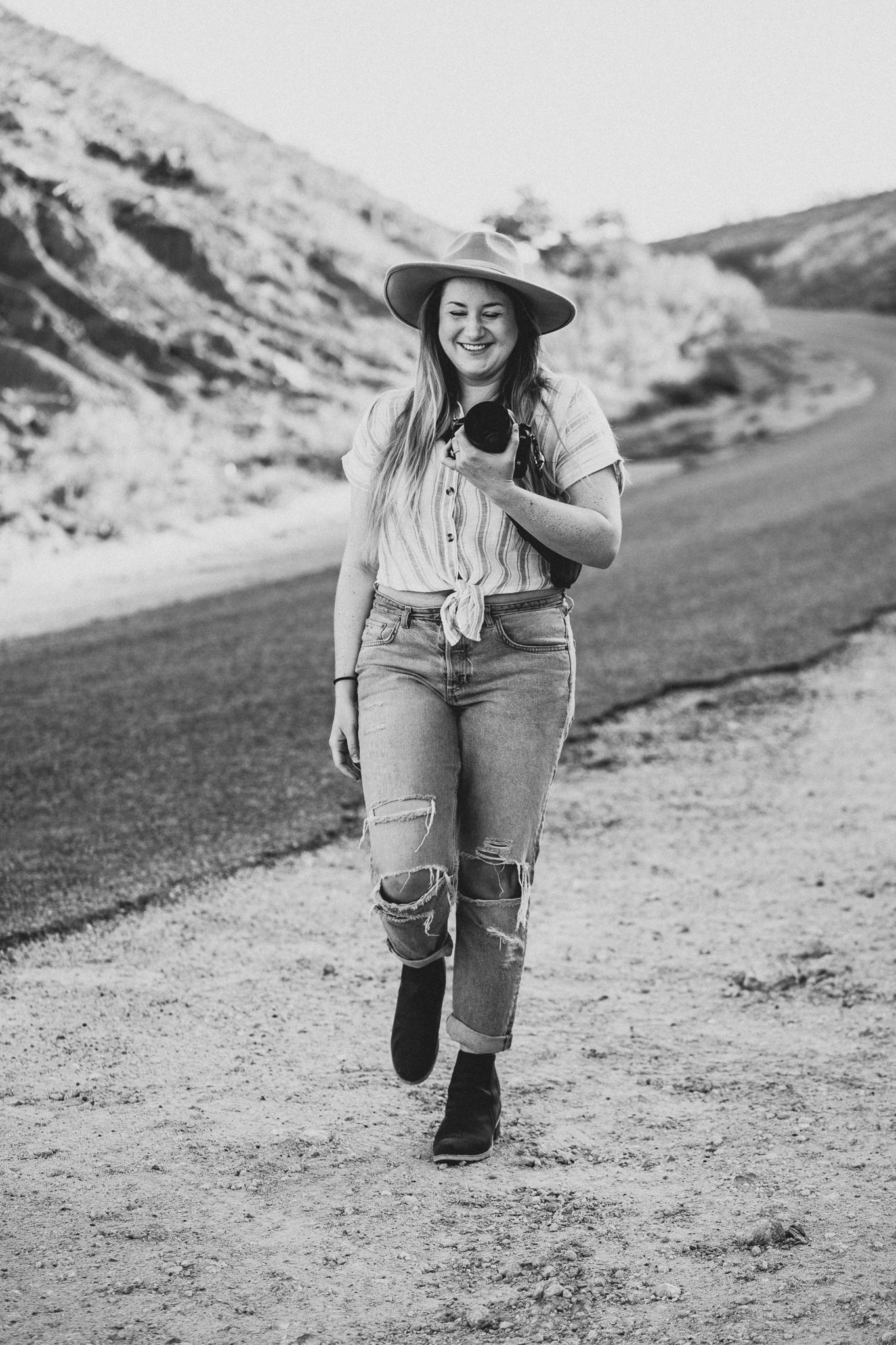 Black and white photo of someone in distressed jeans and boots walking along a dirt path in a mountainous area.