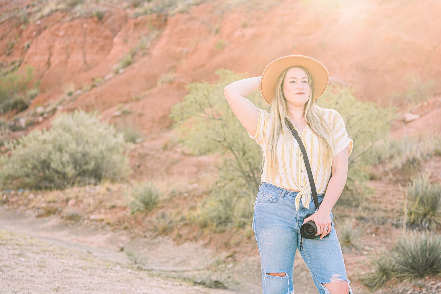 Woman in ripped jeans and white top poses against red rock formations in desert landscape at sunset.
