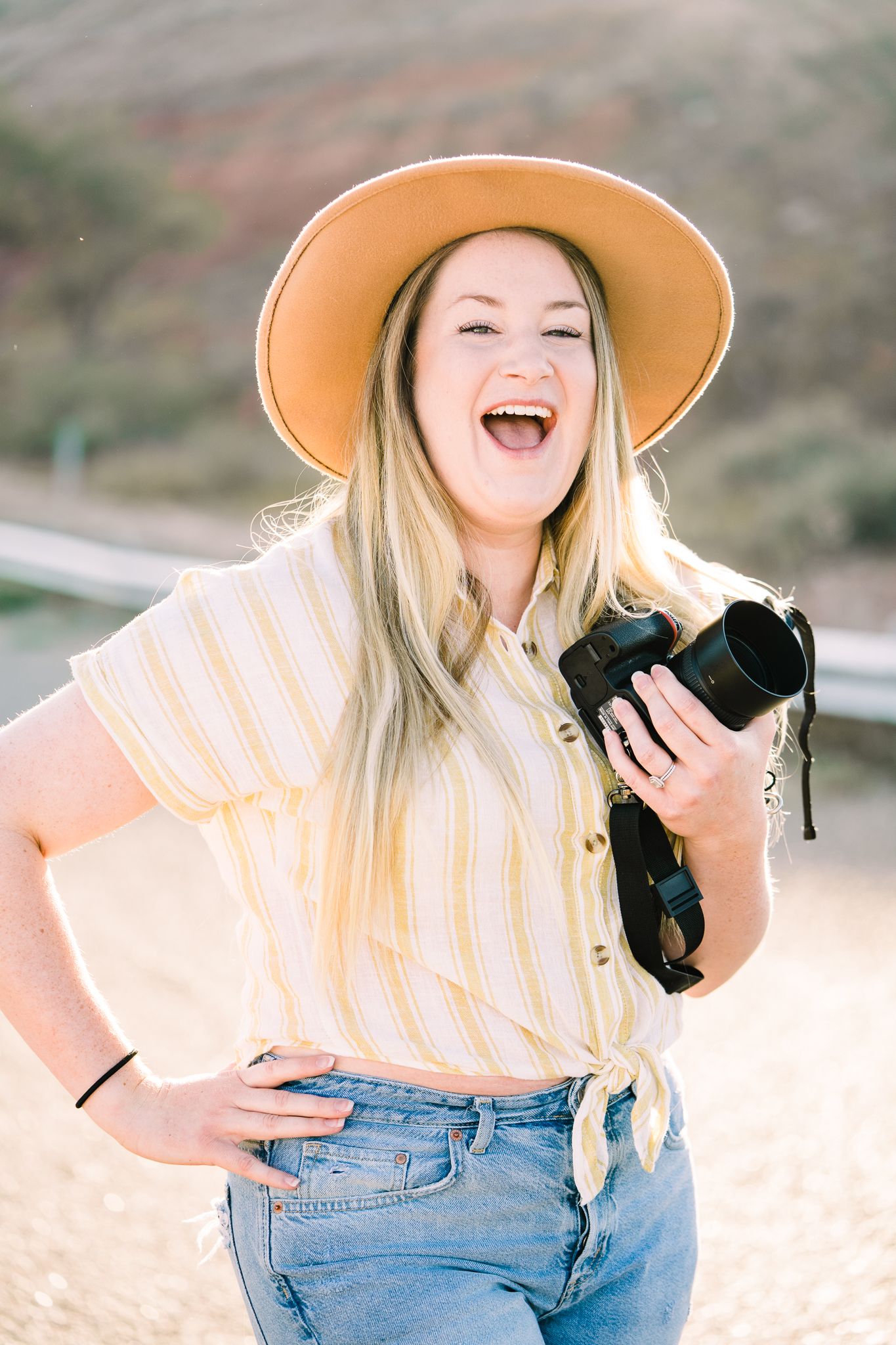 A person in a striped top and denim jeans laughs while holding a black puppy outdoors on a sunny day.
