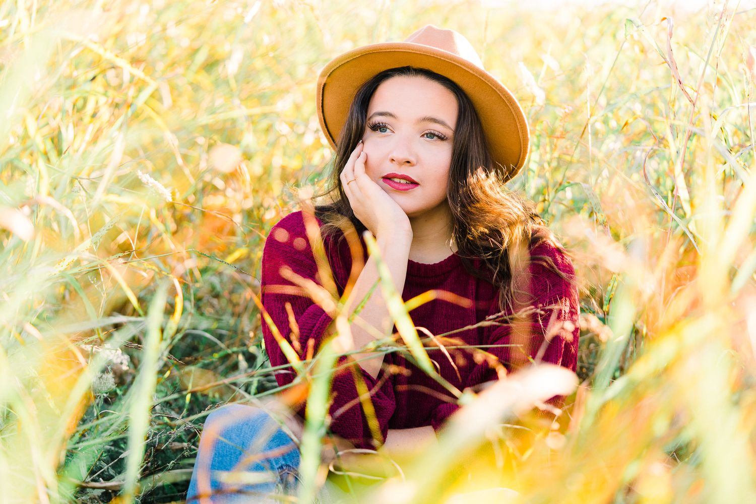 Person in burgundy sweater and brown hat poses joyfully in tall autumn grass during golden hour sunlight.