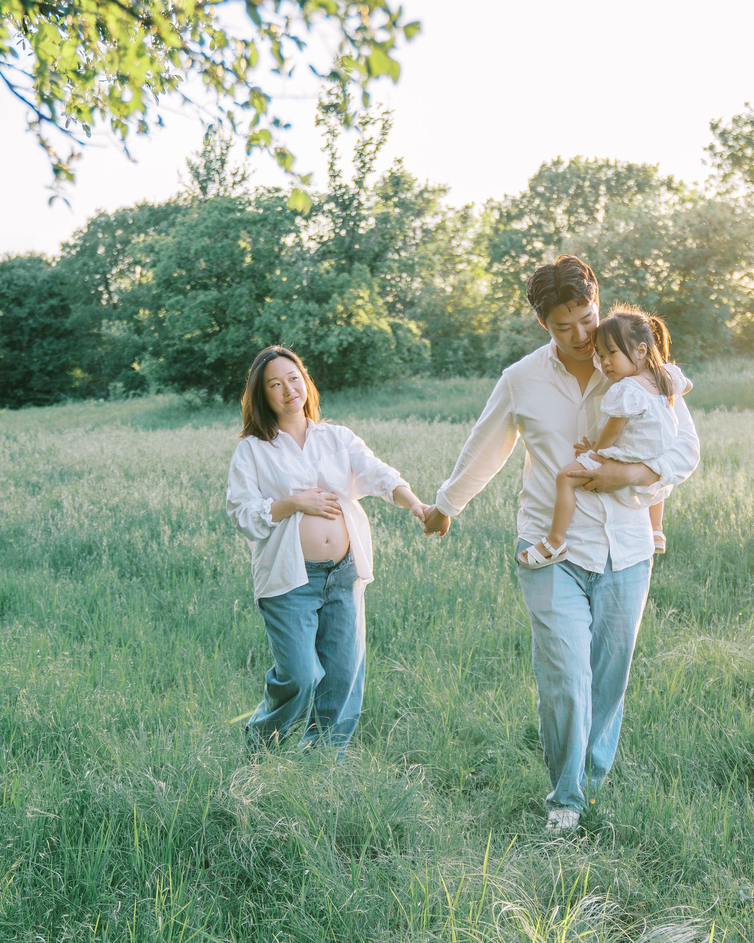 Pregnant woman and man holding hands with toddler in sunny green meadow during golden hour maternity photo session.
