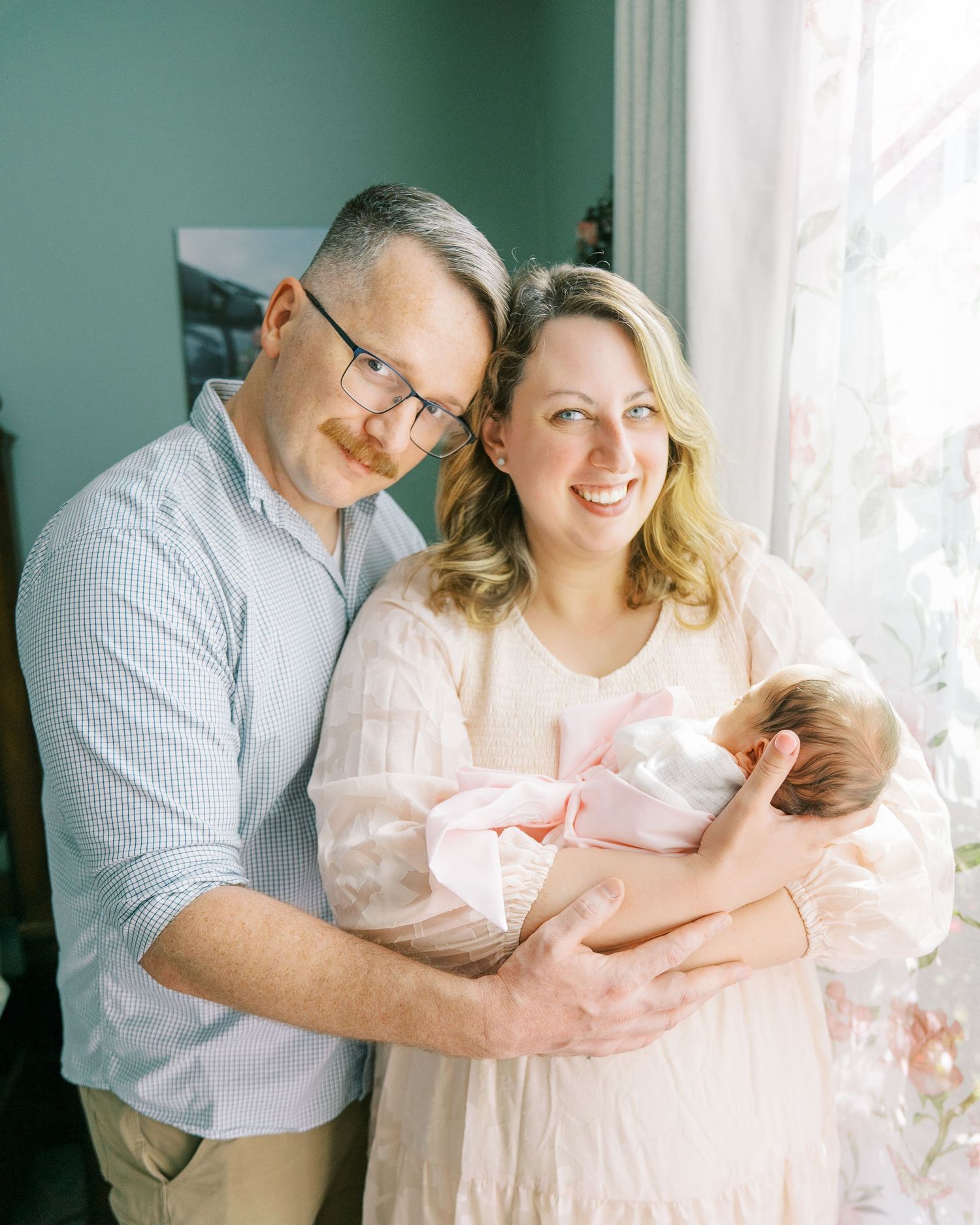 Sweet family moment by window with sage green walls as parents hold their newborn baby.