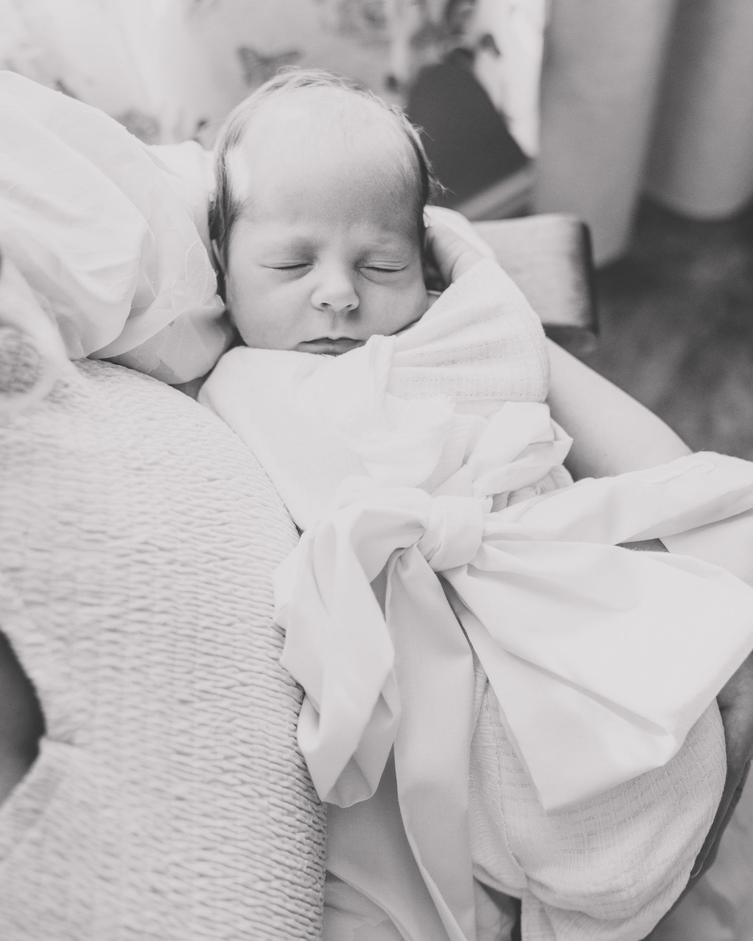 A peaceful sleeping baby swaddled in white blankets rests against a wicker surface in a black and white photograph.