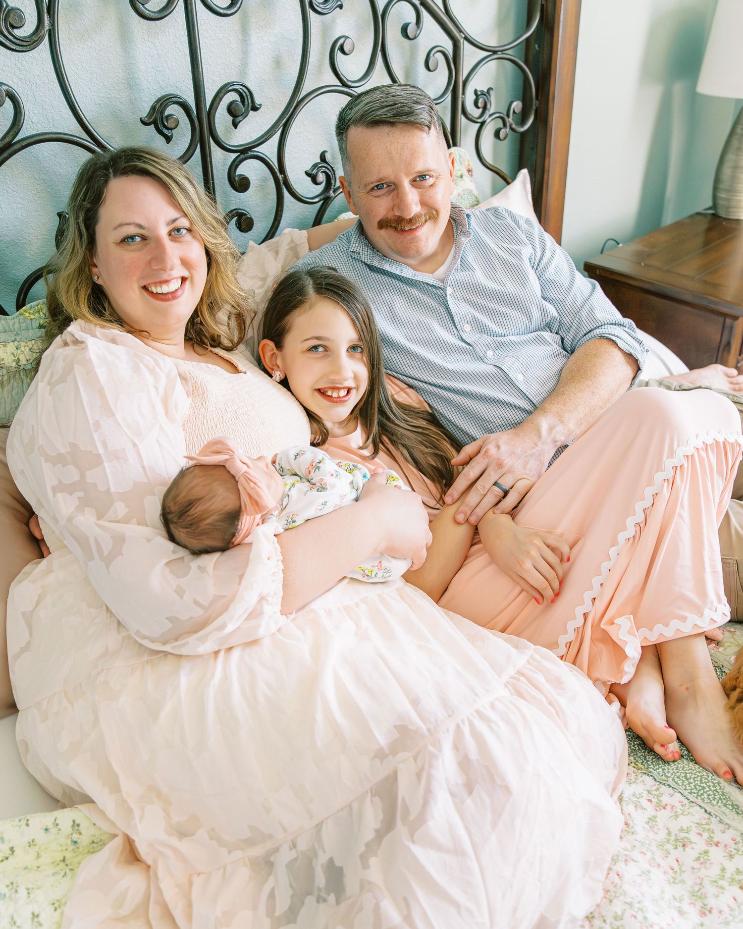 Family lounging together on an ornate bed frame sharing joyful moments with their newborn.