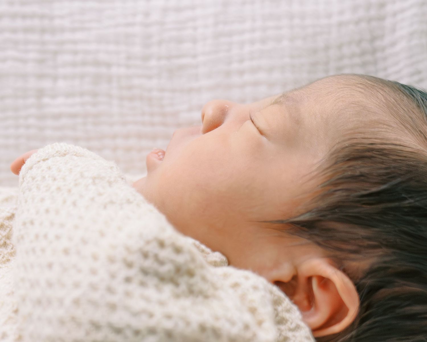Peaceful newborn baby sleeping soundly, wrapped in a soft white knit blanket with dark hair visible.