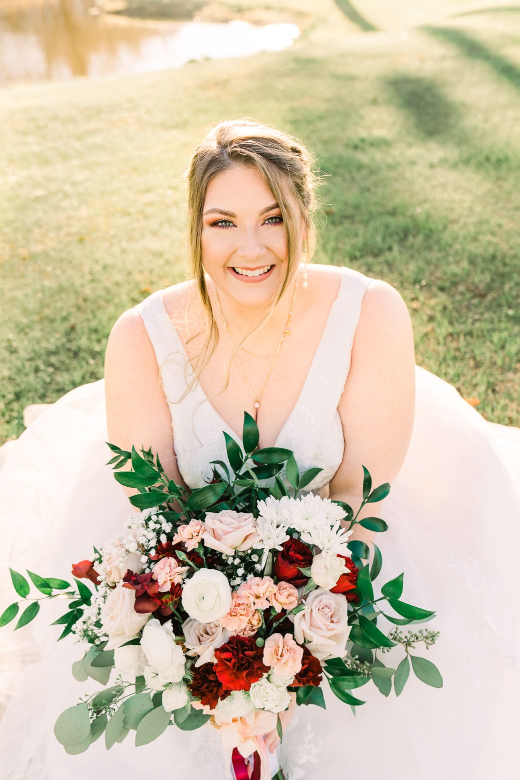 A joyful bride in white dress holds a stunning burgundy and blush bouquet with eucalyptus on a sunny lawn.