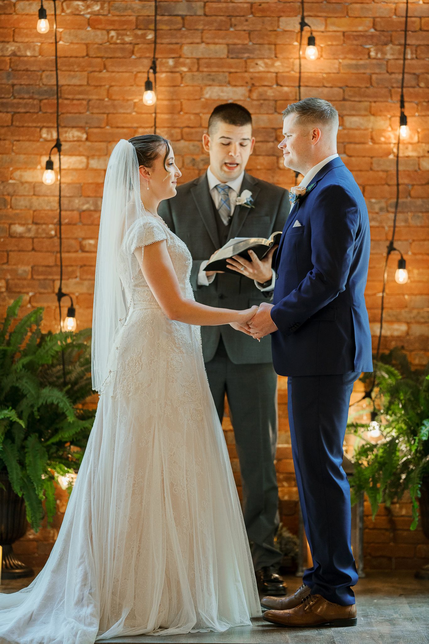 Wedding ceremony in progress with bride in white gown and groom in navy suit against exposed brick wall with hanging lights.