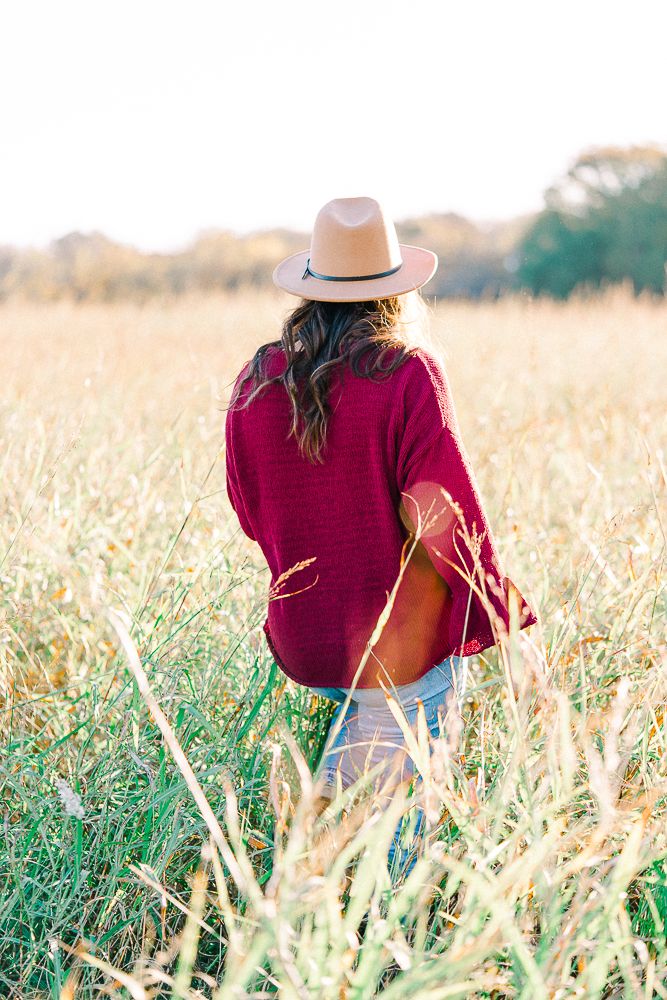 Person in burgundy top and hat sitting in golden wheat field during sunset.