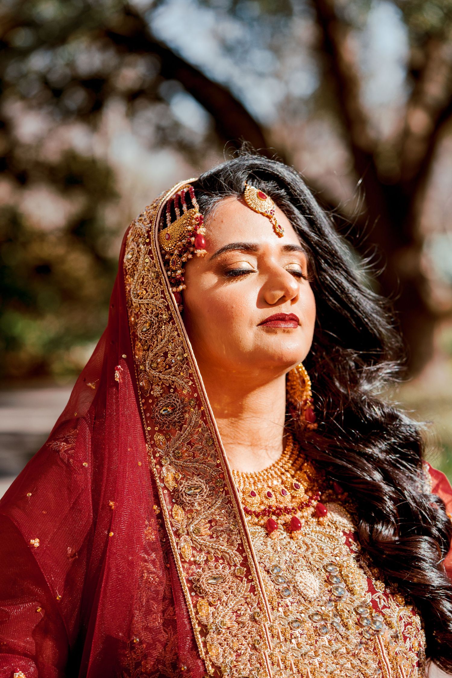Traditional Indian bride in ornate gold and red lehenga poses elegantly with eyes closed in outdoor photoshoot.